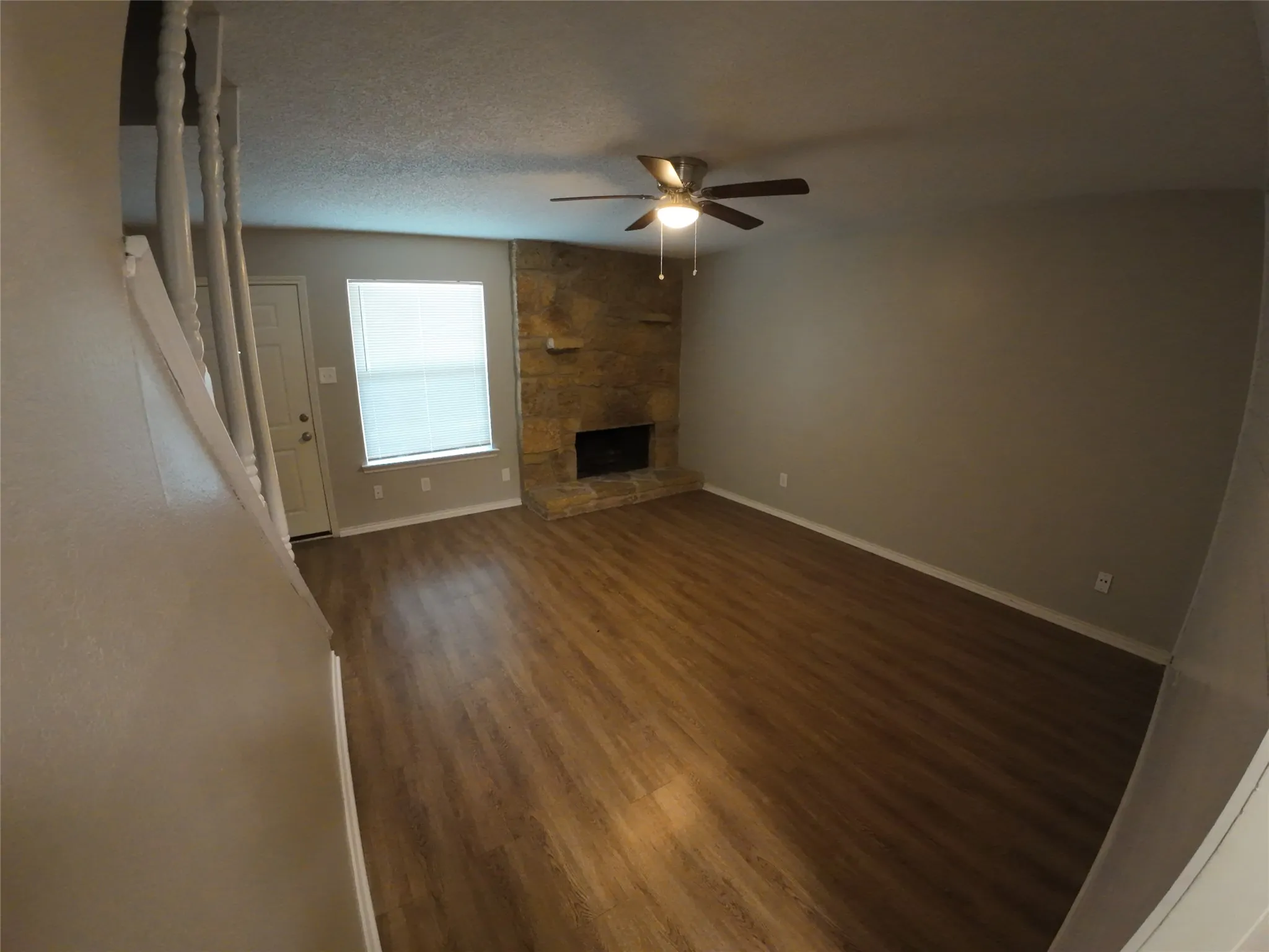 Wood-finish flooring, stone-clad fireplace, ceiling fan with light fixture, and a window with blinds
