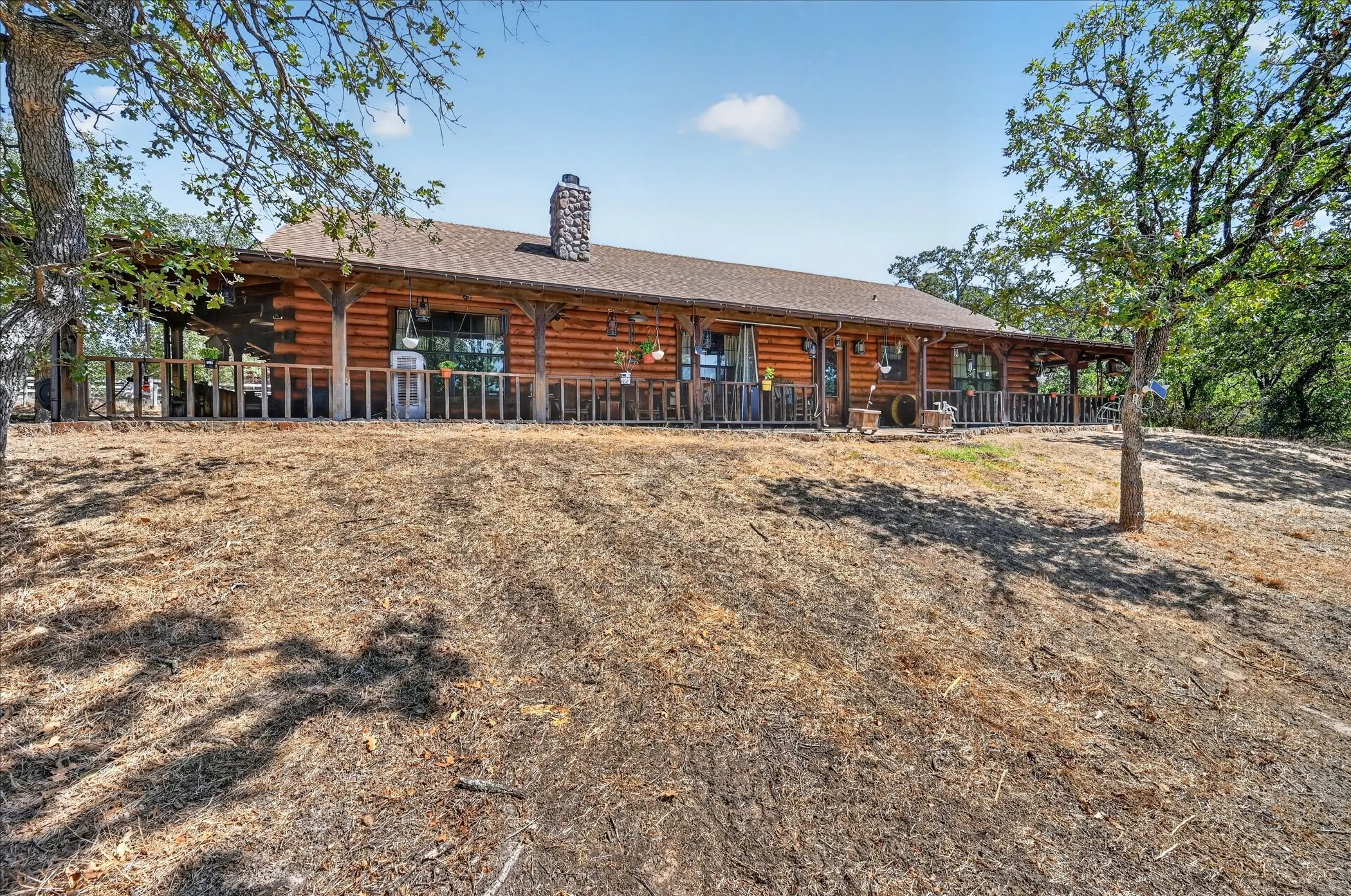 Log cabin exterior with a full-length covered porch, stone chimney, and a sloped terrain