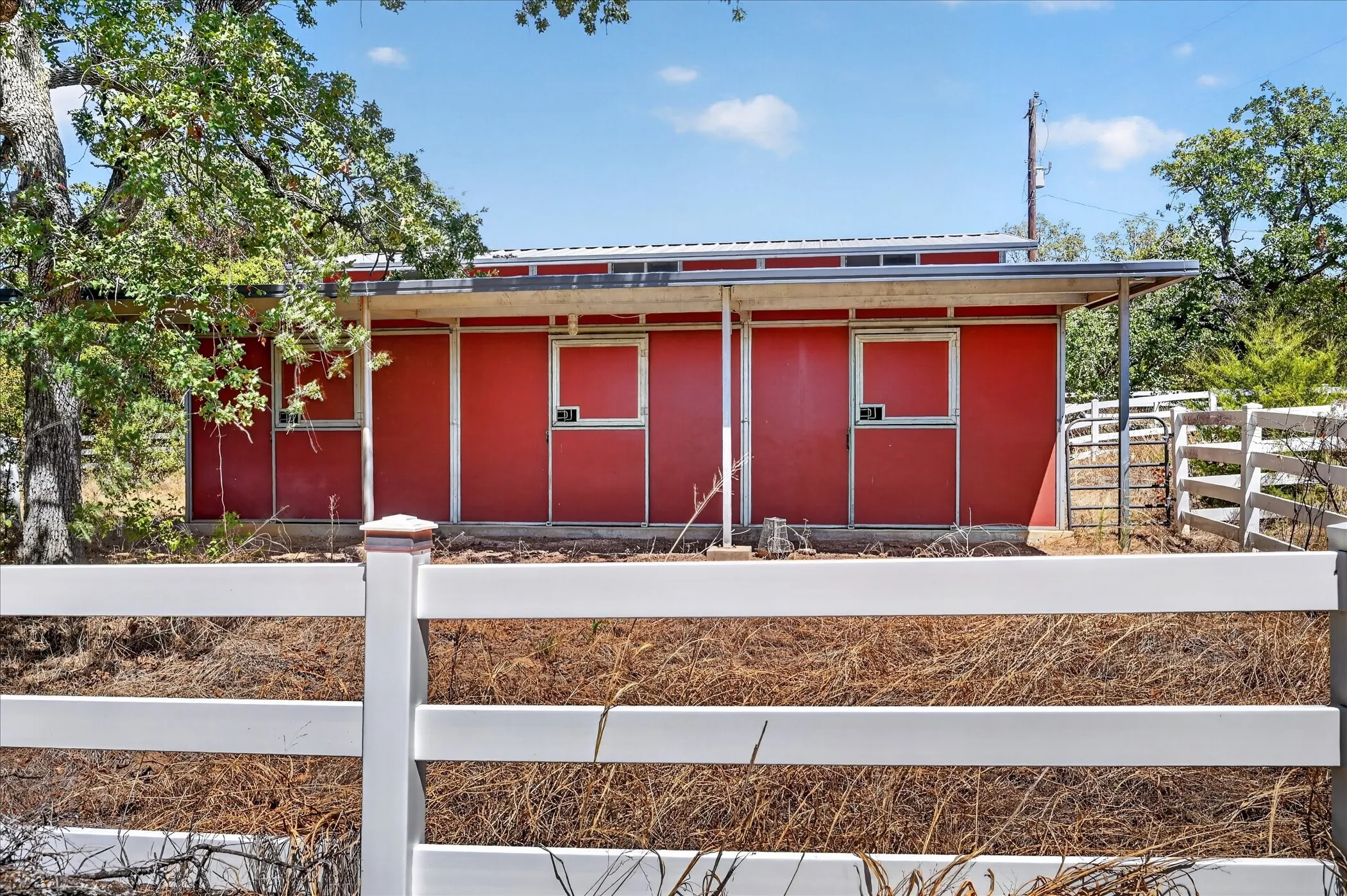 Red barn structure with two stall doors, an extended roofline, and a white fence enclosure