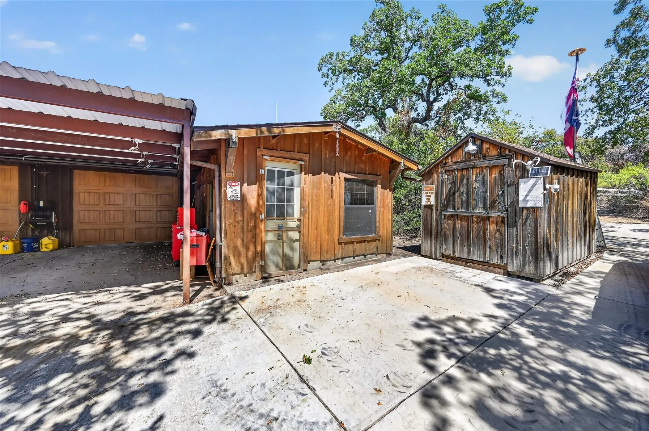 Wood-sided detached structures with a covered parking area featuring a roll-up garage door, a separate building with a paneled door and window, and an additional shed with a solar panel