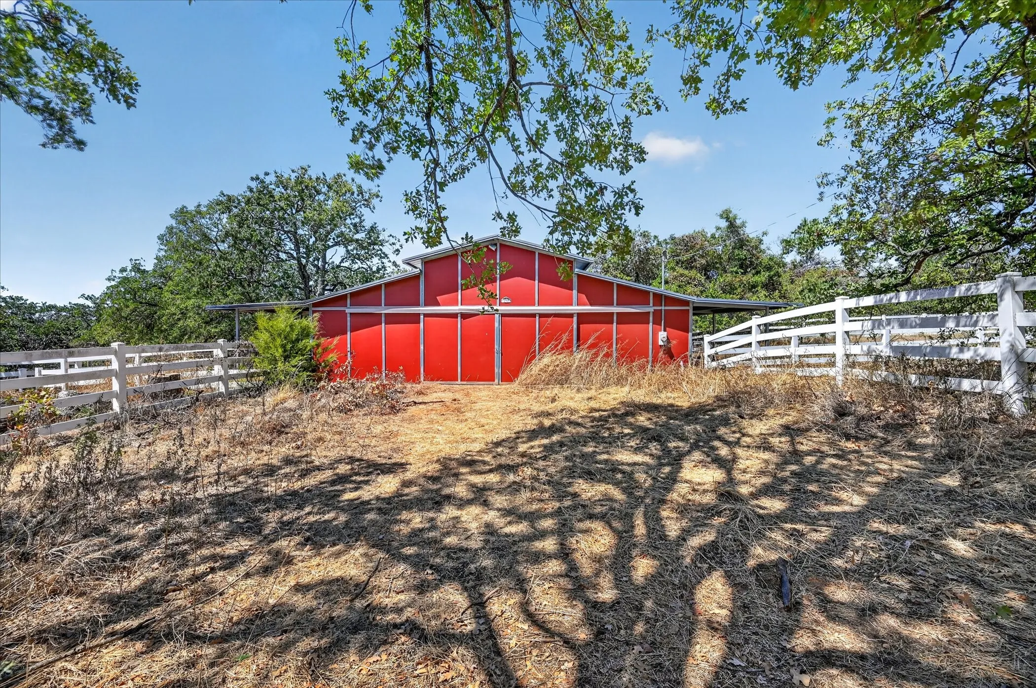 Red barn structure with a metal roof, white perimeter fencing, and mature trees