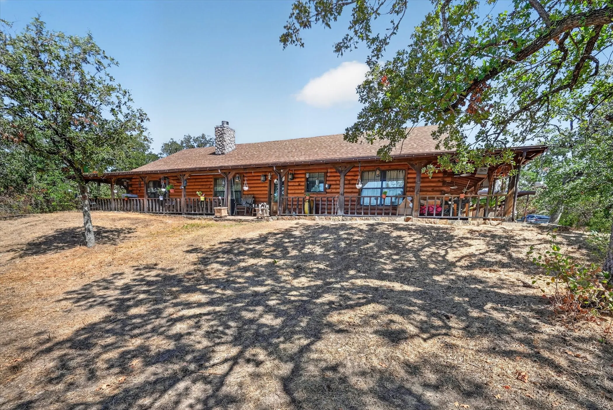 Log cabin exterior with a stone chimney, extensive covered front porch, and surrounding mature trees