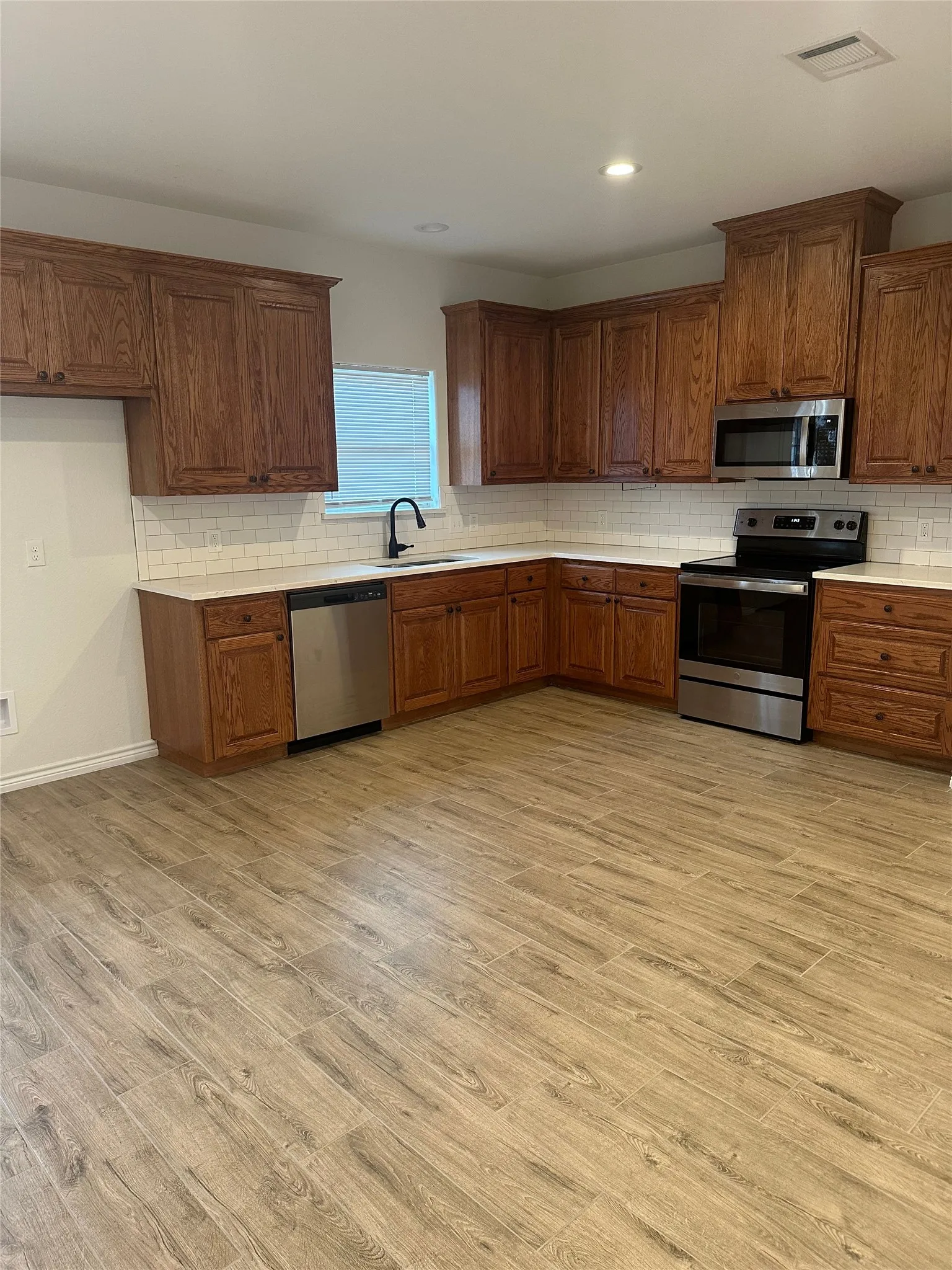 Kitchen with wood-finish flooring, wood cabinetry, stainless steel appliances, white subway tile backsplash, and recessed lighting