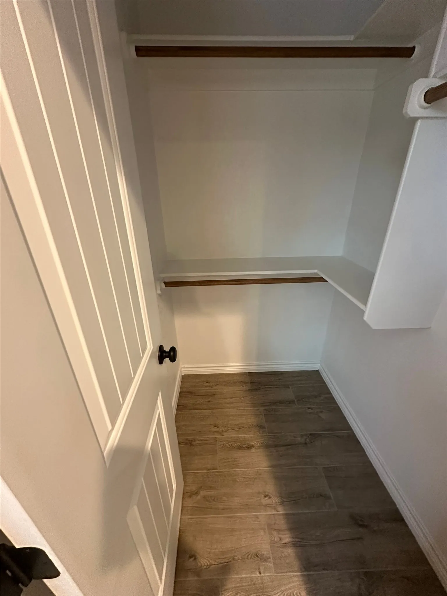 White interior closet with wood-finish flooring, two wood-tone hanging rods, and a white paneled door with a black knob