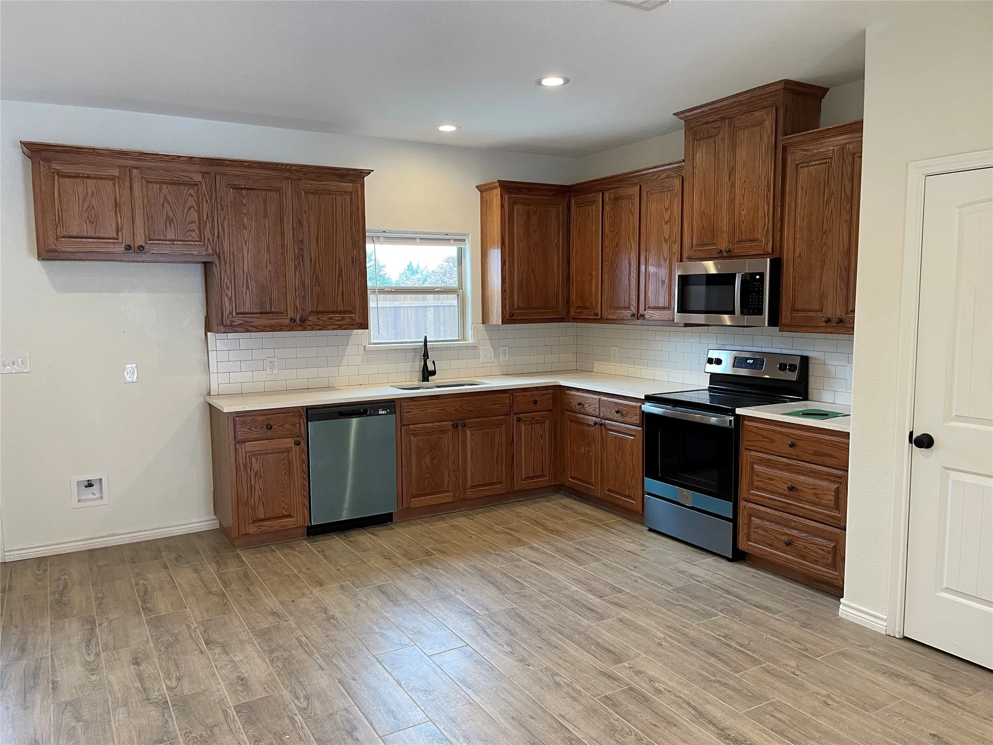 Kitchen with wood-finish cabinetry, stainless steel appliances, white subway tile backsplash, and light-toned countertops
