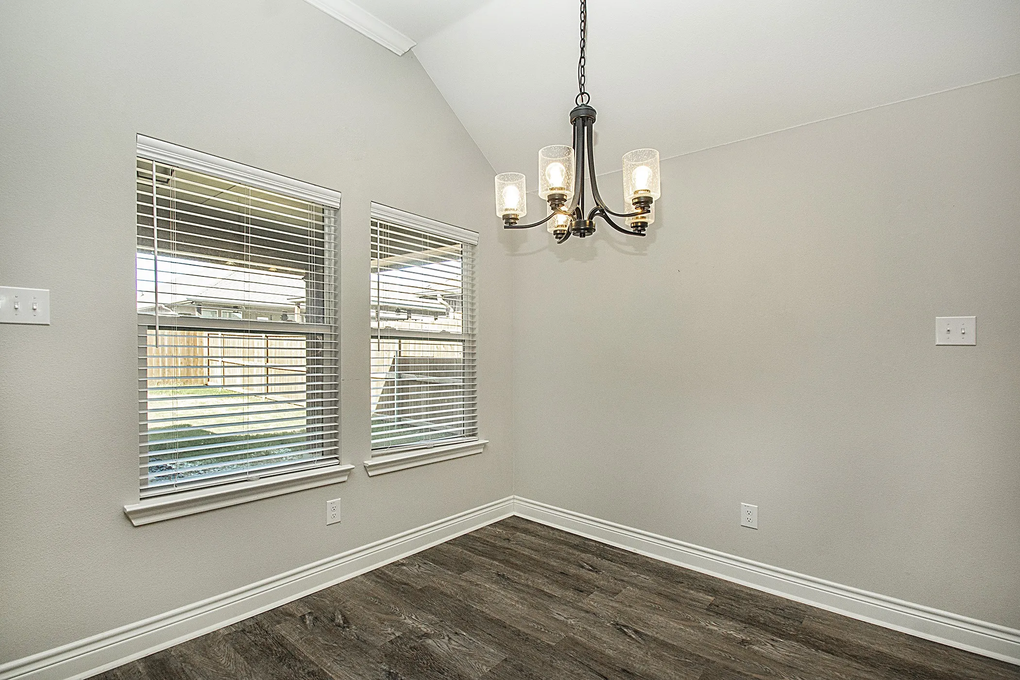 Vaulted ceiling, four-light chandelier, two windows with blinds, wood-finish flooring, and white baseboards