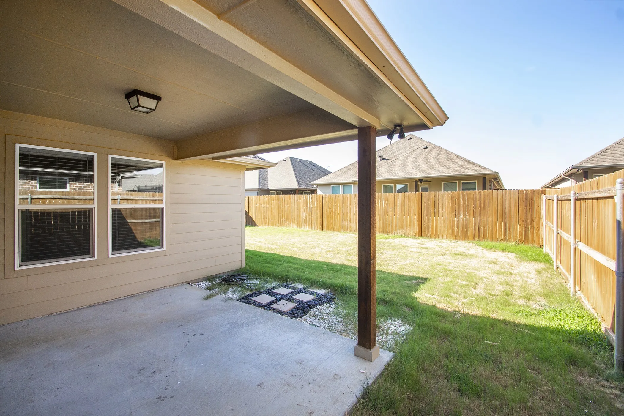 Covered concrete patio with an outdoor light fixture, wood support column, and a fully fenced backyard featuring a grassy area
