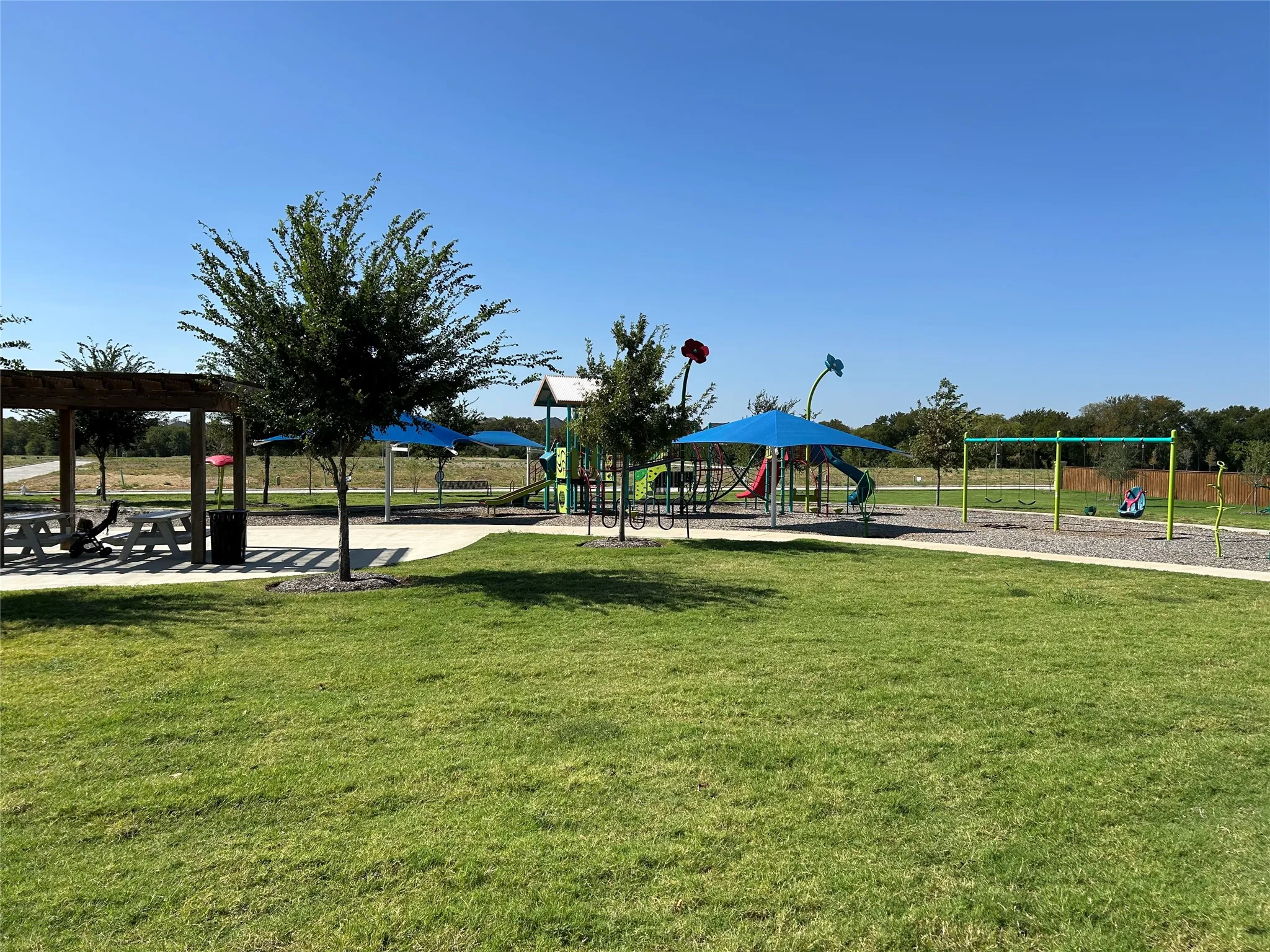 Expansive green space featuring playground equipment, swings, shaded seating areas, and a paved pathway