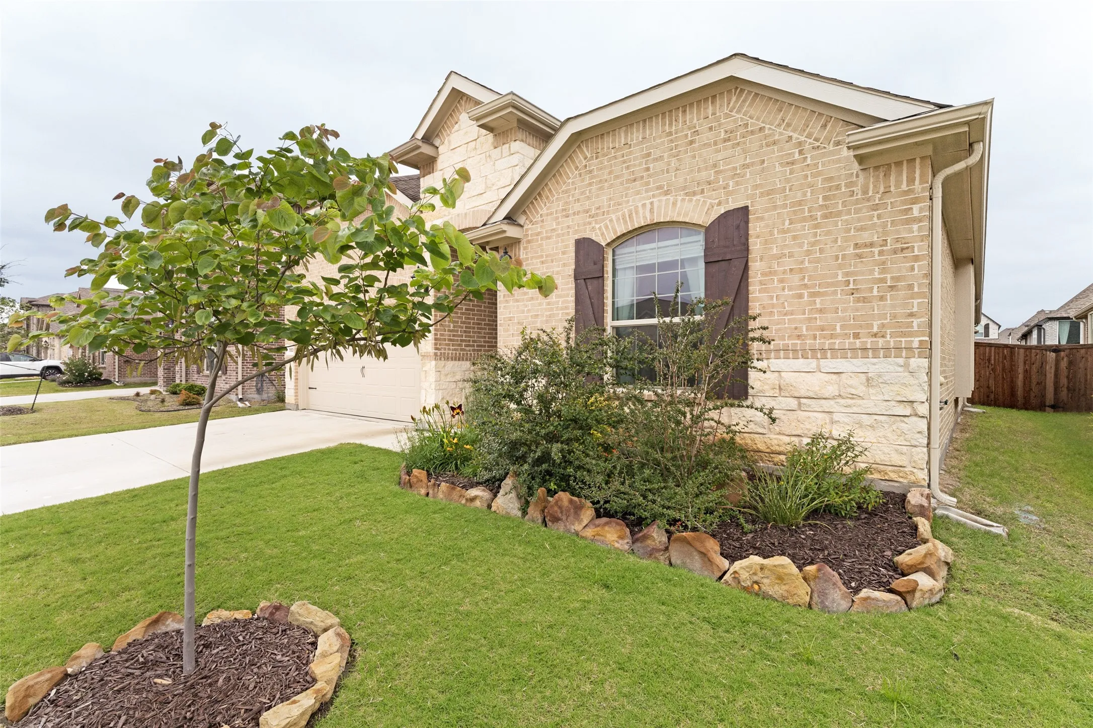 Brick and stone exterior with decorative window shutters, arched window, attached garage, and a landscaped front yard with a young tree