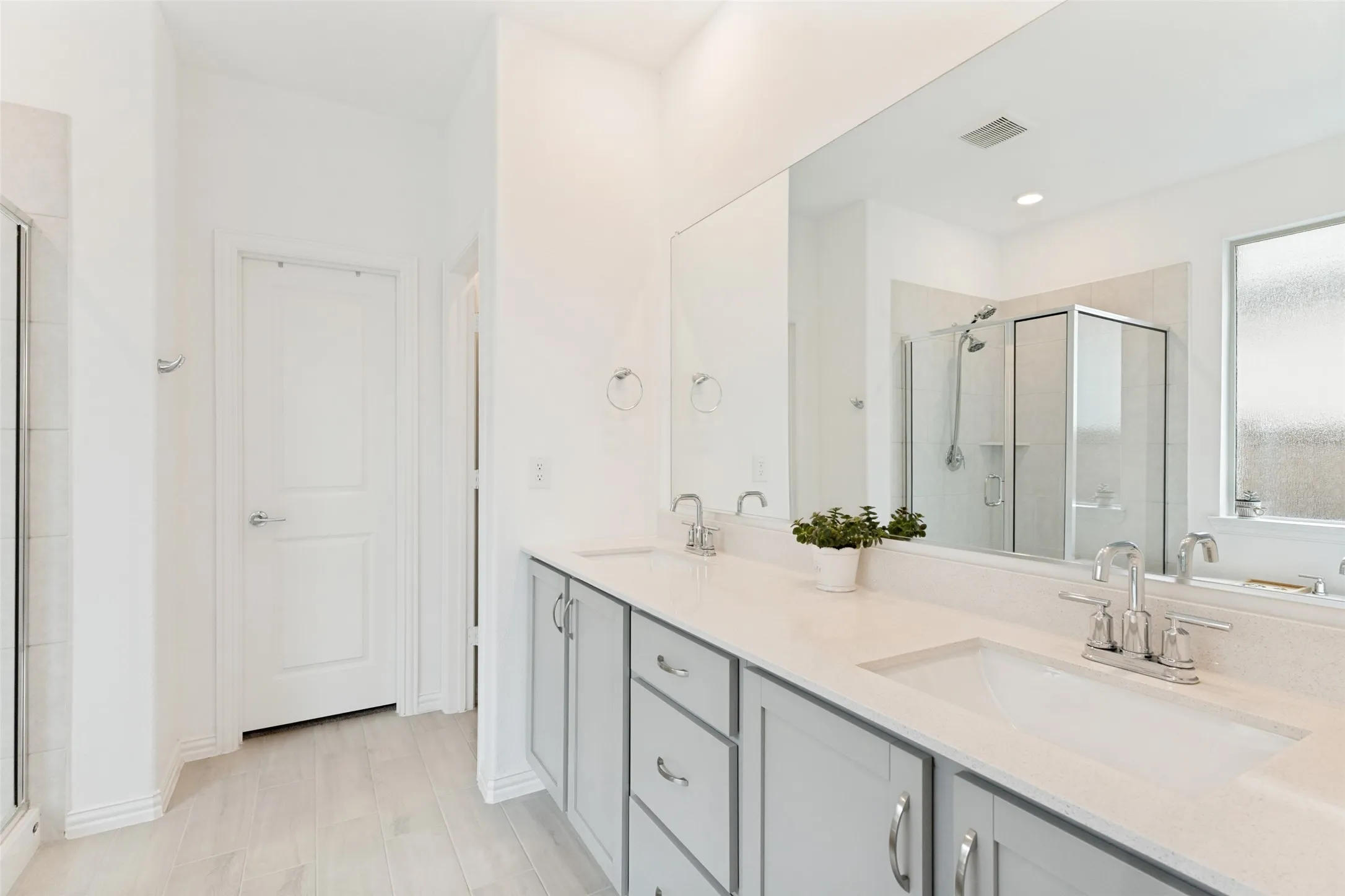 Dual vanity with integrated sinks, light wood-finish flooring, glass-enclosed shower, and a large wall-mounted mirror