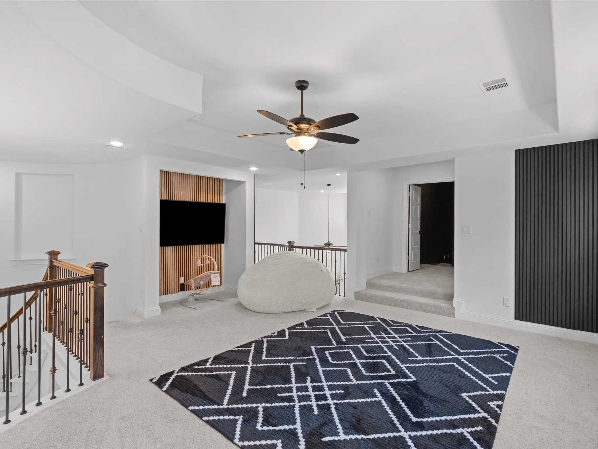 Spacious carpeted area with a ceiling fan, recessed lighting, and a dark wood and iron railing