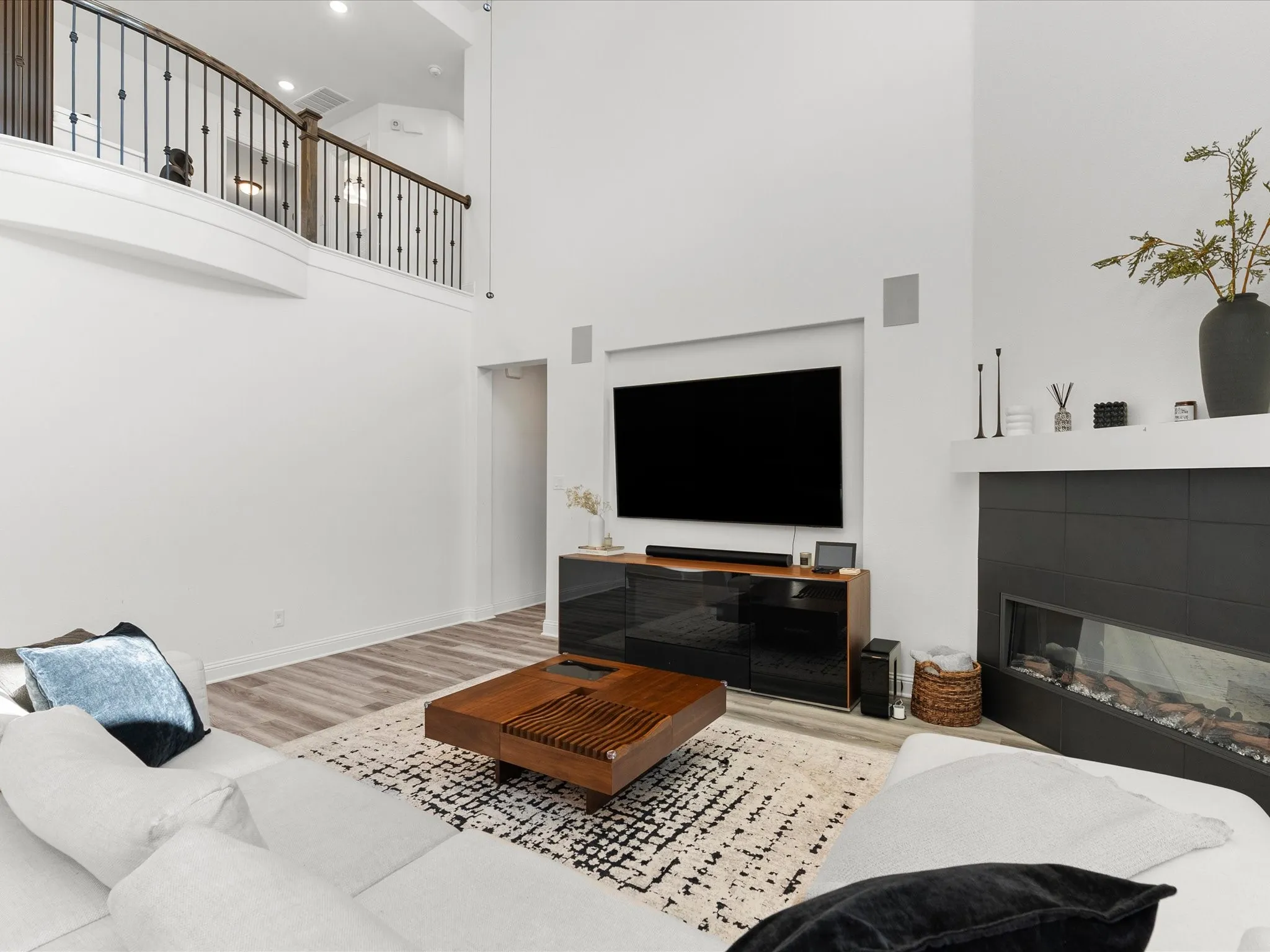 Expansive two-story living space featuring high ceilings, wood-finish flooring, a contemporary fireplace with black tile surround, and a wrought iron and wood-finish railing