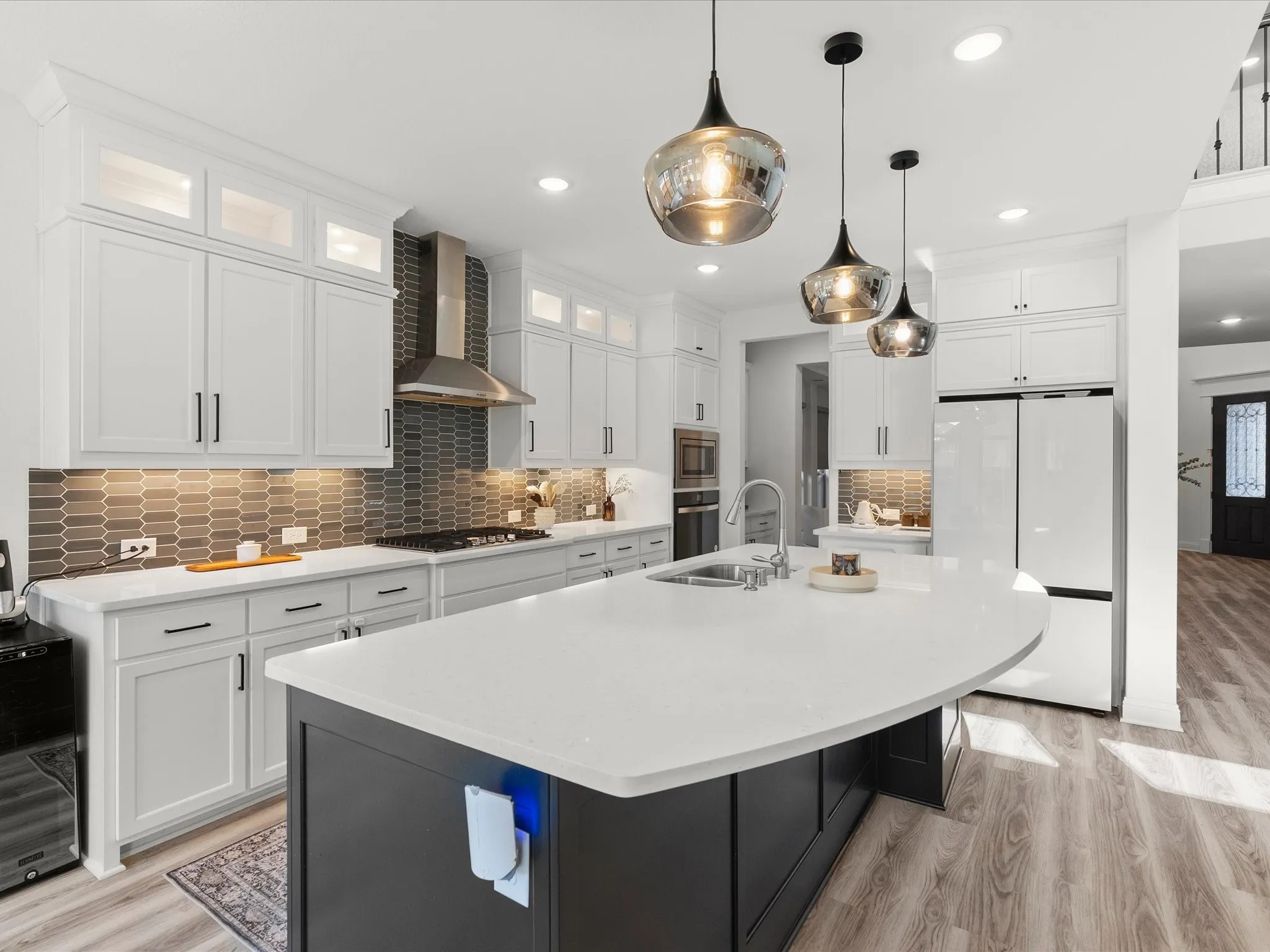 White shaker cabinetry with upper glass-front cabinets, stone countertops, gray geometric tile backsplash, and a large kitchen island with pendant lighting