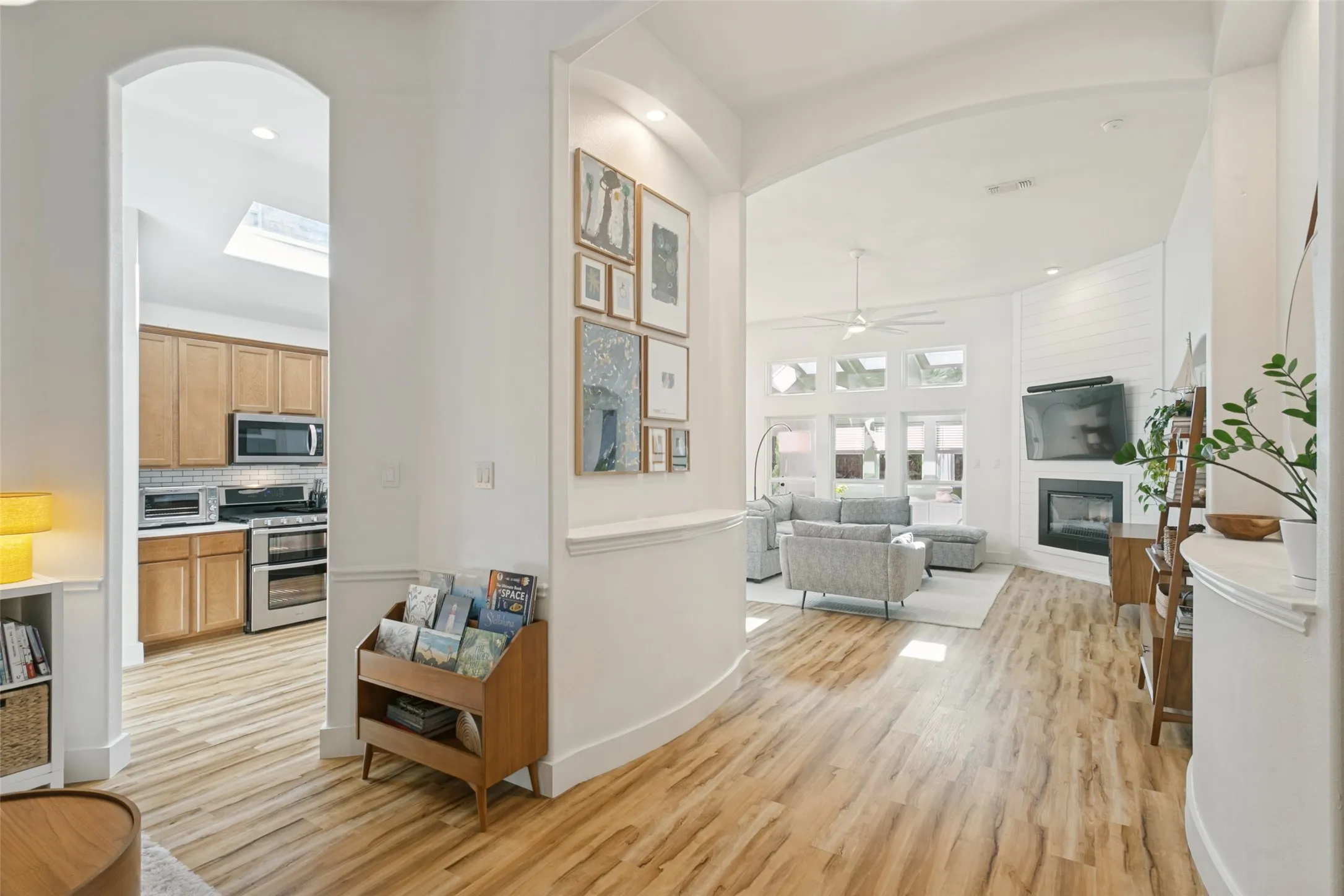 Wood-finish flooring throughout, arched entryway, a kitchen with wood cabinetry and stainless steel appliances, a living area with a ceiling fan, and a fireplace with a mantel
