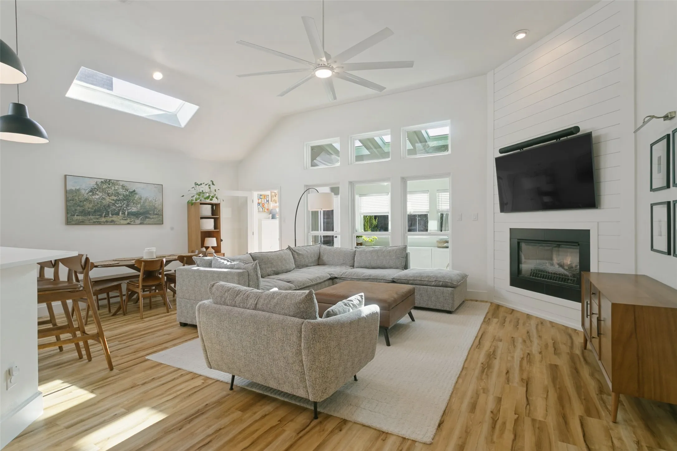 Vaulted ceiling with skylight, wood-finish flooring, white shiplap fireplace surround, recessed lighting, and a ceiling fan
