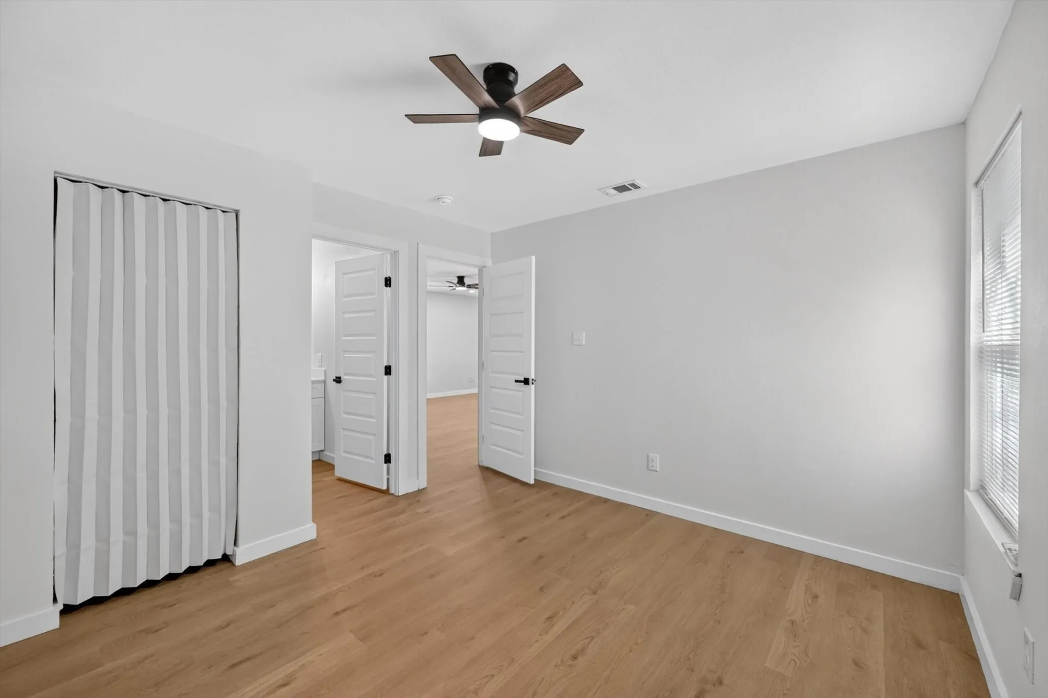 Bright secondary bedroom featuring gleaming wood-finish flooring, soft neutral wall finishes, a ceiling fan with integrated lighting, and multiple closet spaces with white panel doors