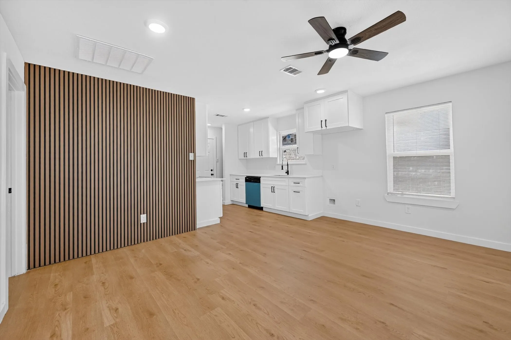Open-concept living space featuring warm wood-finish flooring, white shaker-style cabinetry with integrated appliances, ceiling fan with brushed metal finish, recessed lighting, and a striking vertical slat accent wall in natural wood tones