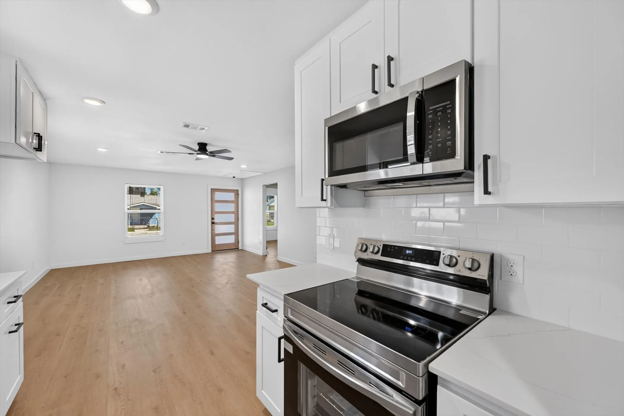 Stainless steel electric range and microwave, white shaker cabinetry with matte black hardware, white subway tile backsplash, light wood-finish flooring, and a modern ceiling fan