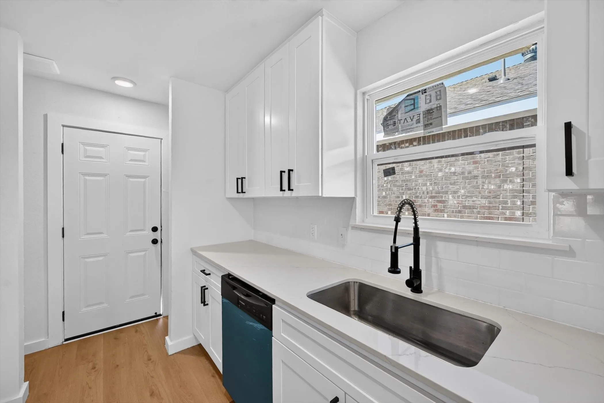 White shaker cabinetry with matte black hardware, light-toned countertops, subway tile backsplash, stainless steel sink with a modern faucet, and wood-finish flooring