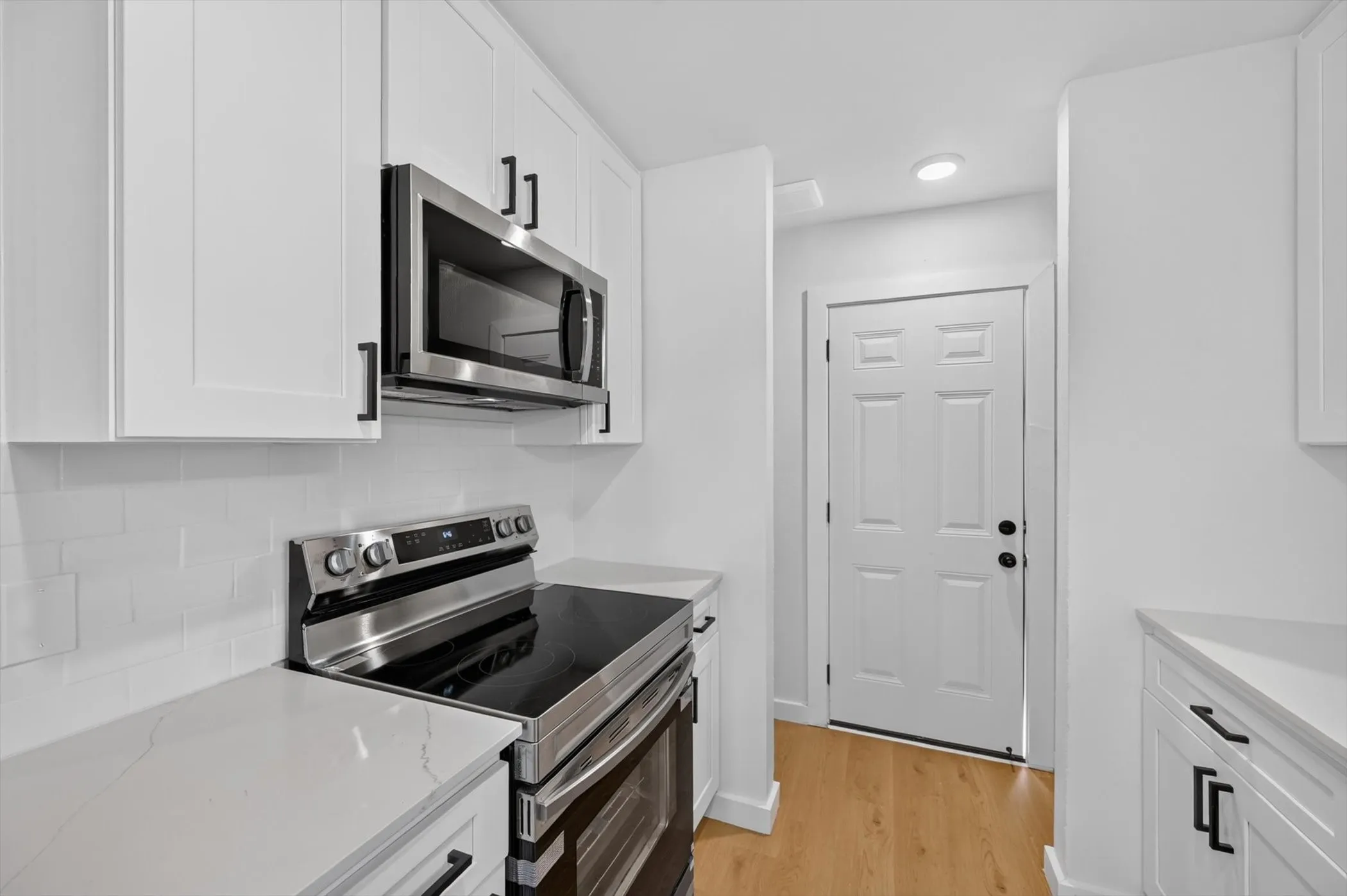 White shaker cabinetry with matte black hardware, stainless steel appliances, white subway tile backsplash, light-toned wood-finish flooring, and white paneled door