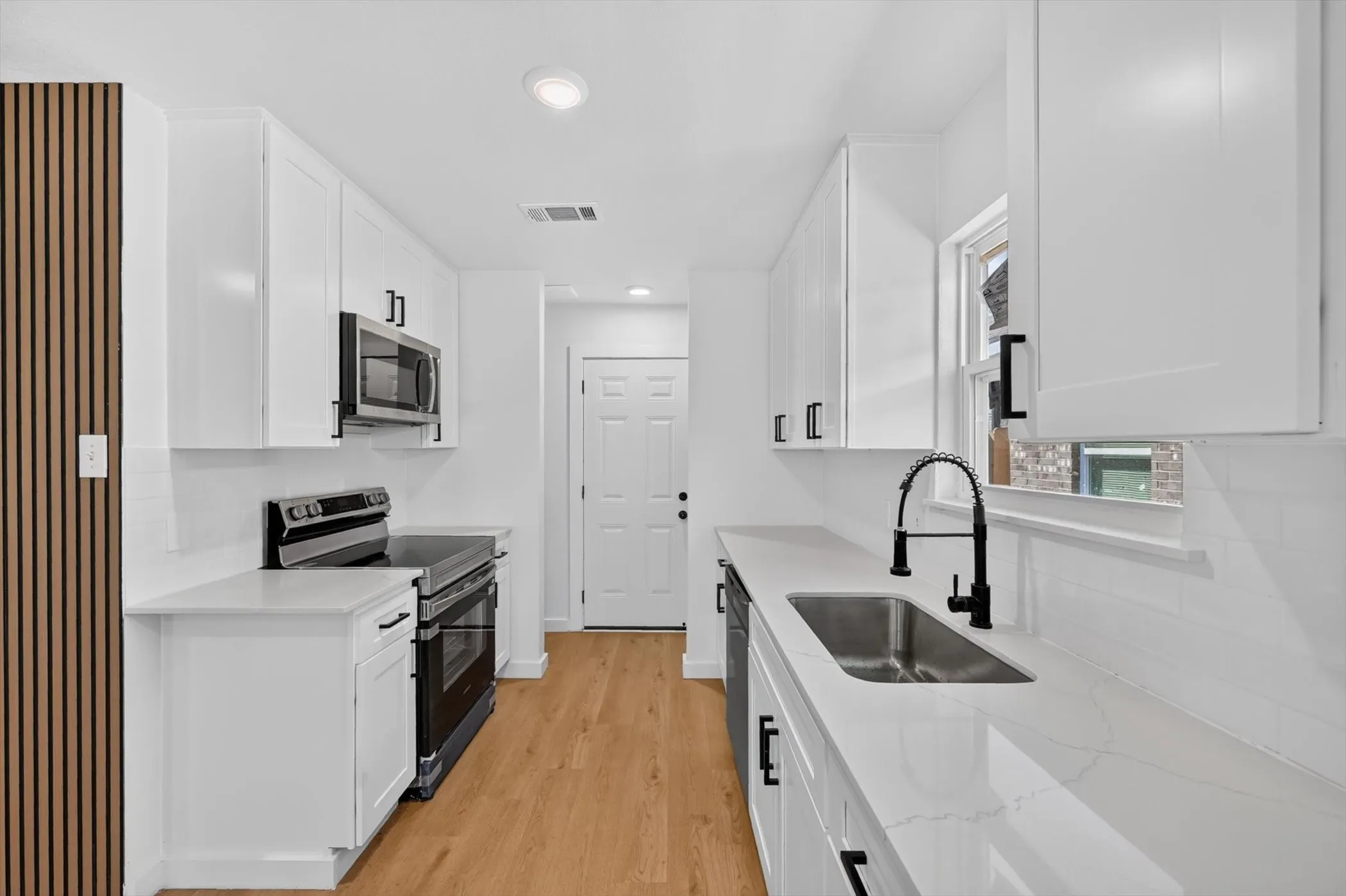 Modern kitchen featuring white shaker cabinets, light wood-finish flooring, stainless steel appliances, a deep basin sink with a matte black faucet, and white countertops