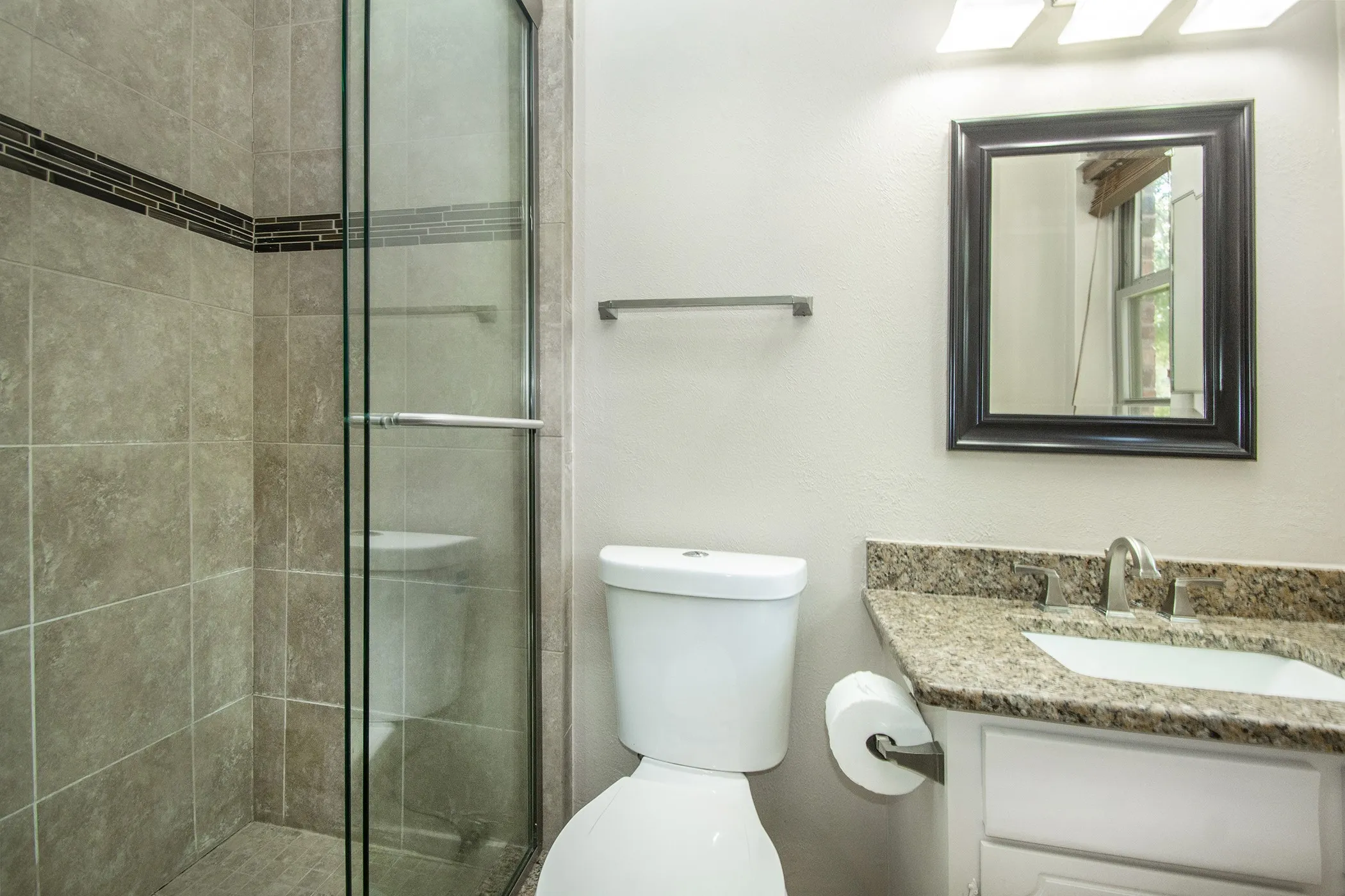 Glass-enclosed shower with tile surround and decorative accent band, vanity with stone countertop, undermount sink, and wall-mounted mirror