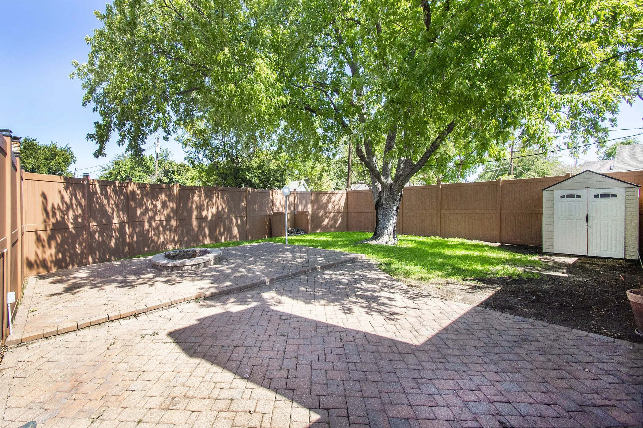 Expansive backyard featuring a paver patio, stone fire pit, mature tree, and a storage shed