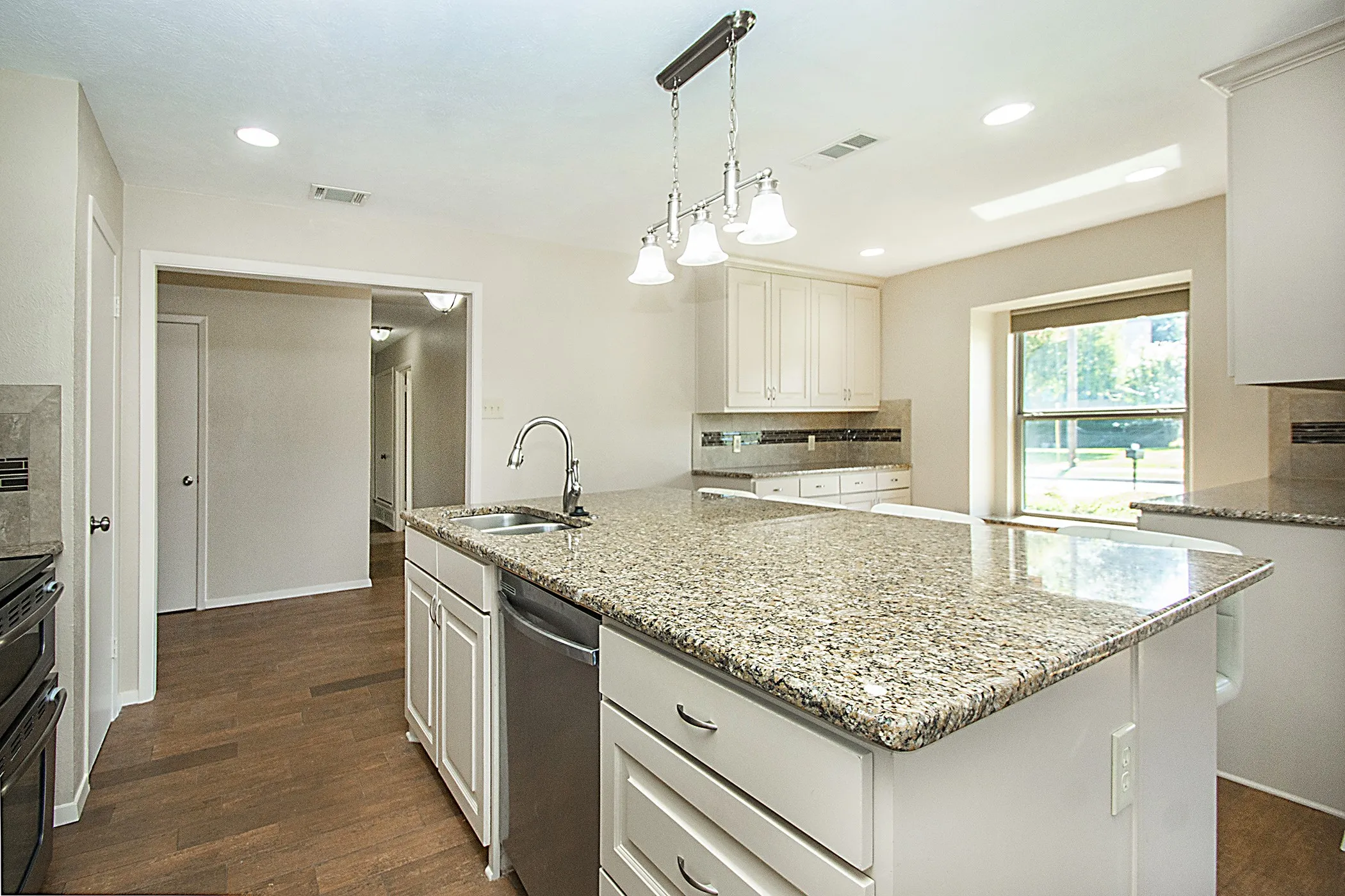 Kitchen featuring an island with a built-in sink, stainless steel dishwasher, white cabinetry, granite countertops, and wood-finish flooring