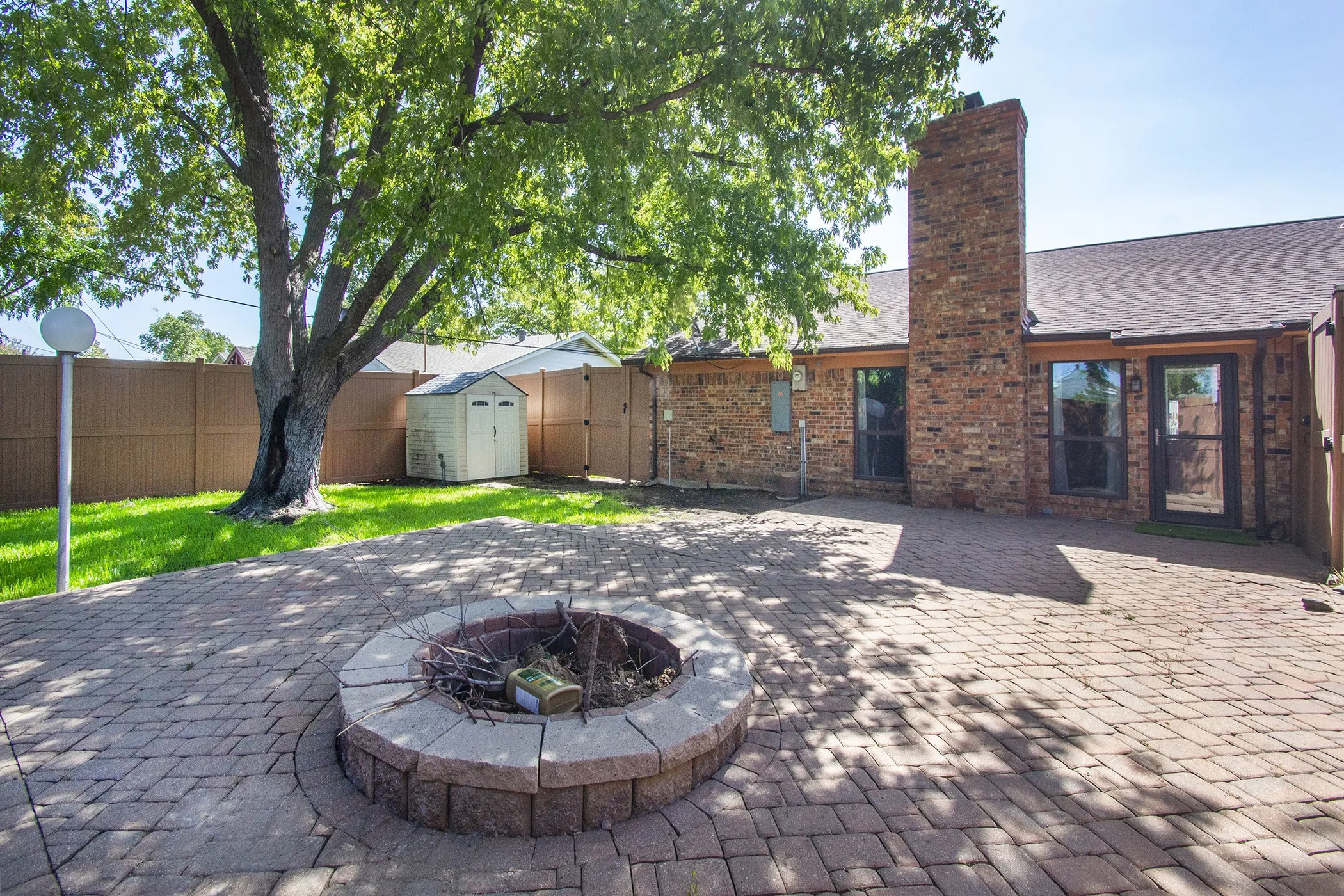 Expansive paver patio featuring a built-in fire pit, mature shade tree, and brick exterior
