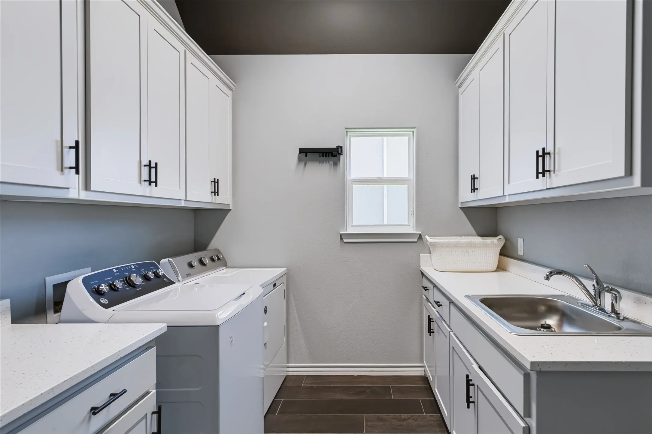 White shaker-style cabinetry with black hardware, light-toned countertops, stainless steel utility sink with chrome faucet, wood-finish flooring, and a single window
