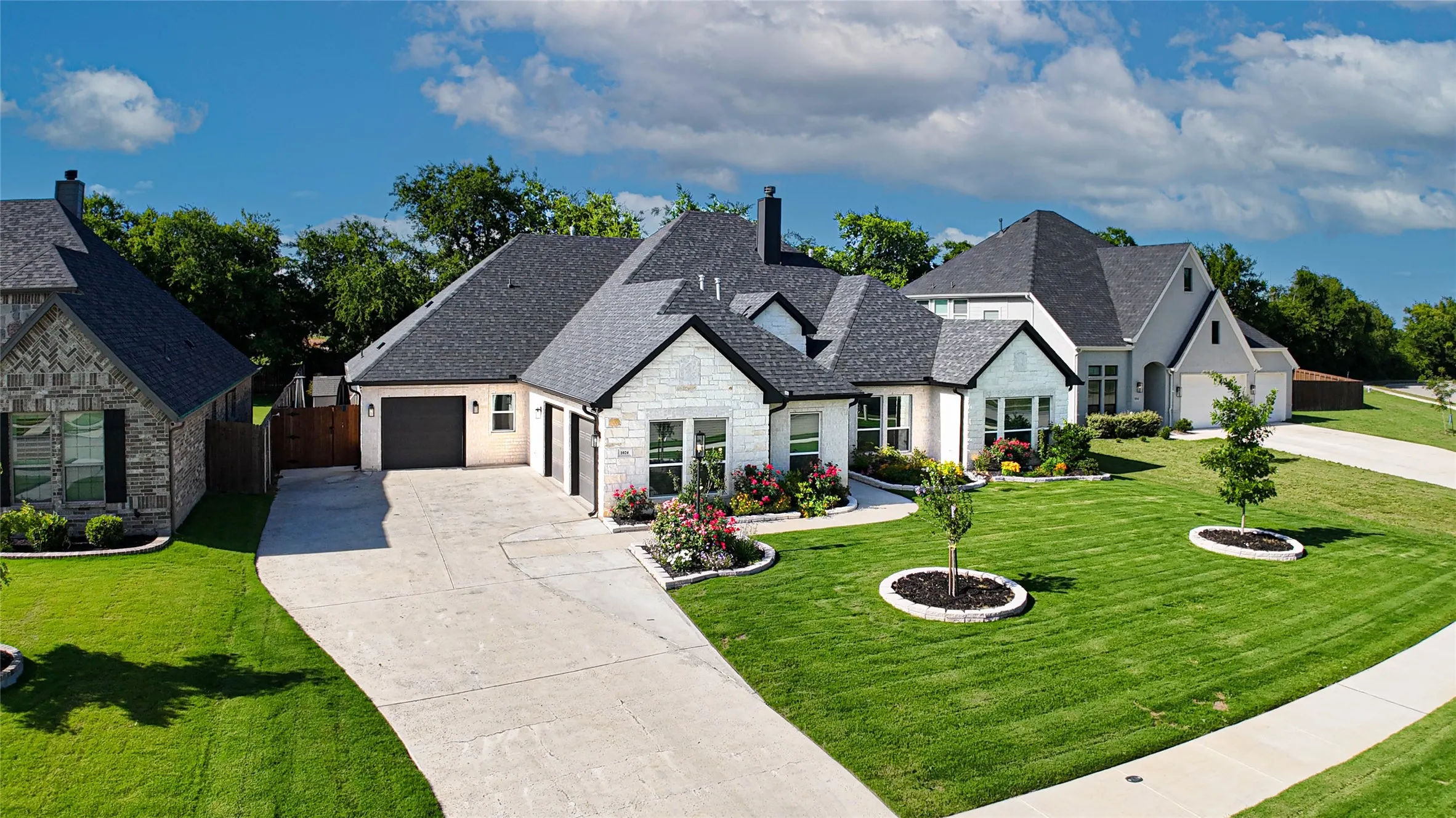 White stone and stucco exterior with dark trim accents, concrete driveway, attached garage, well-maintained lawn, and established landscaping