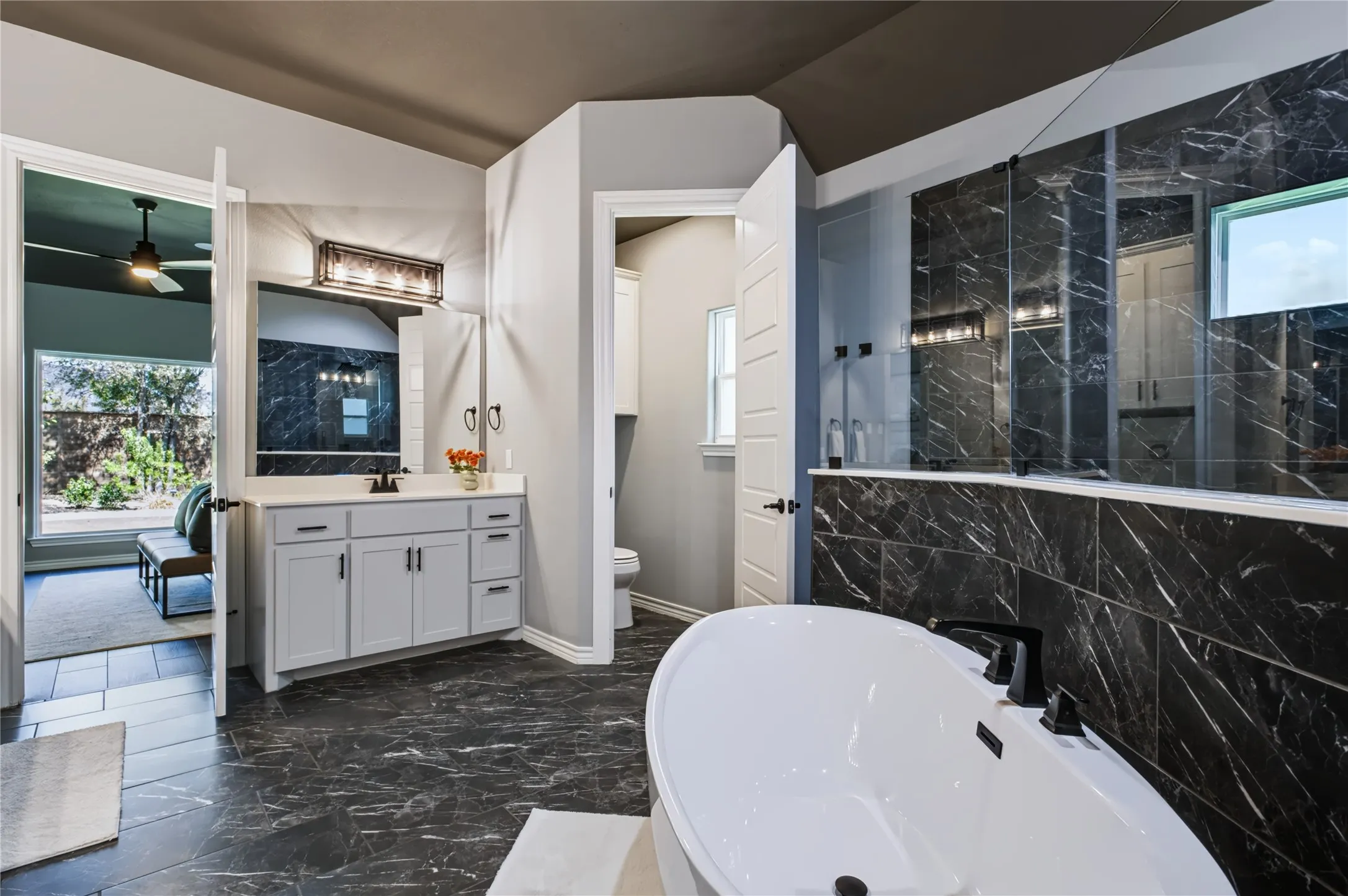Freestanding soaking tub with black fixtures, dark marble-look tile flooring, white vanity with dual sinks, and an expansive walk-in shower with dark marble-look tile