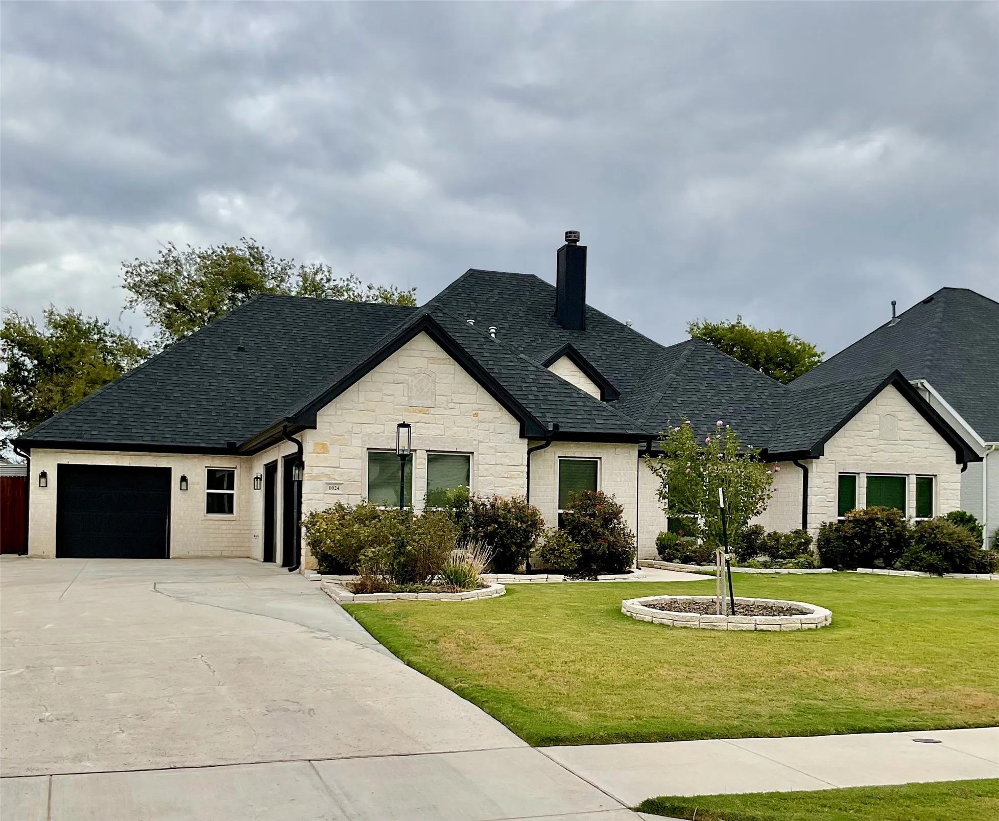 White brick exterior with dark trim, black garage door, dark shingle roof, and a meticulously landscaped front yard with stone-edged planters