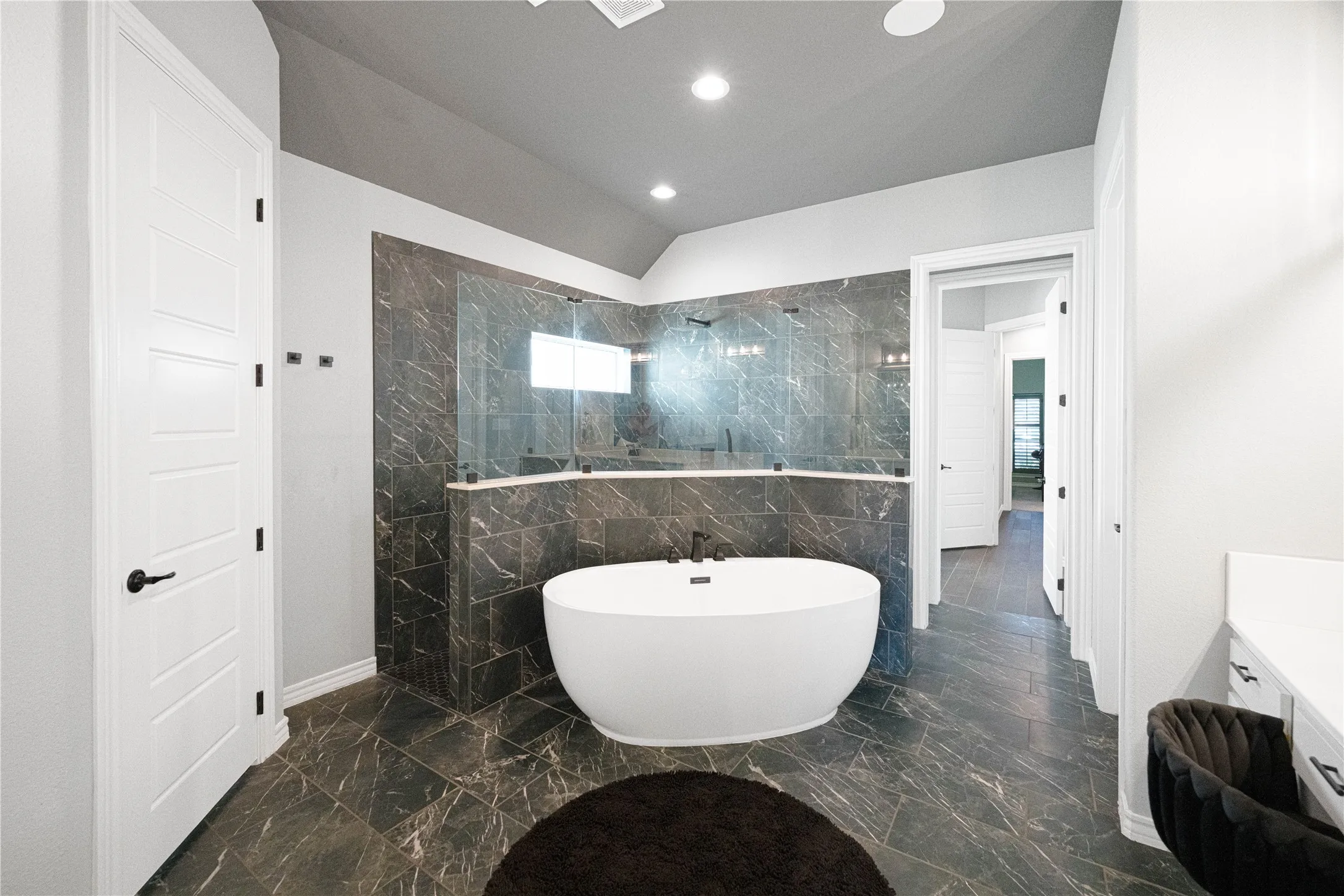 Freestanding soaking tub, large glass-enclosed shower, dark marble-finish tile flooring, vaulted ceiling, and recessed lighting