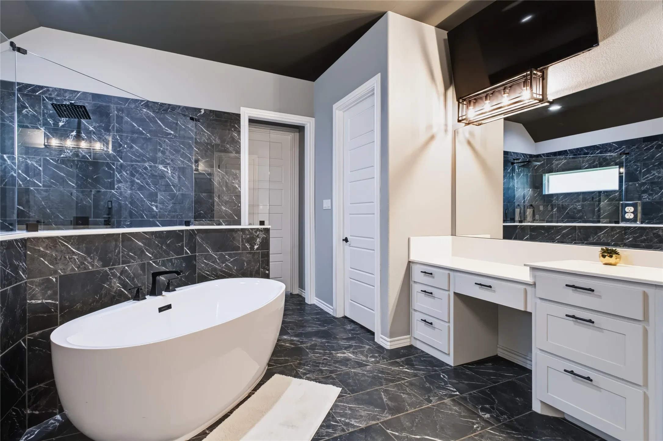 Freestanding soaking tub, black-tiled shower with glass enclosure, dual vanity with white countertops, built-in makeup vanity, and large format floor tiles