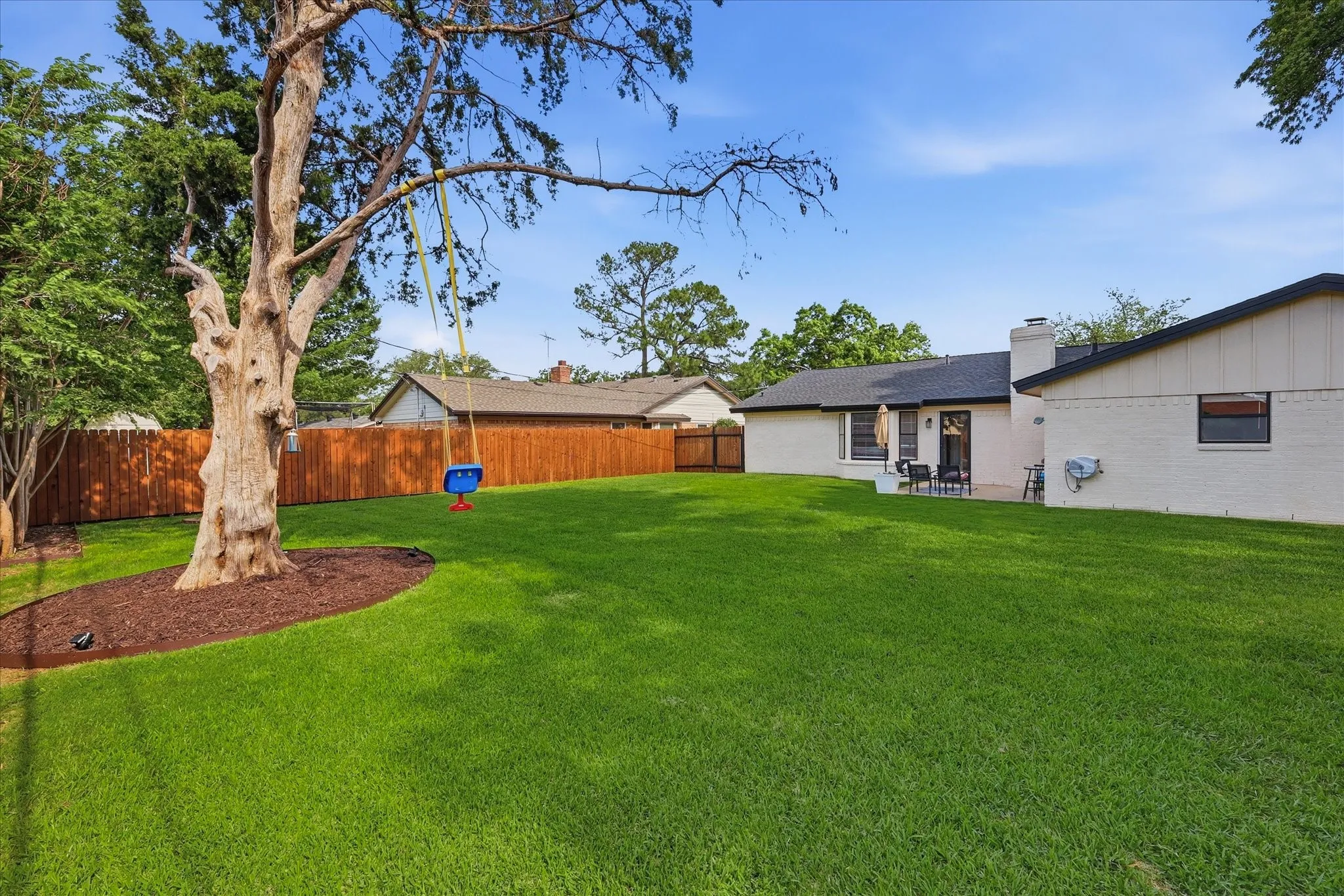 Expansive green lawn, mature trees, wood privacy fencing, patio area with outdoor seating, and brick exterior