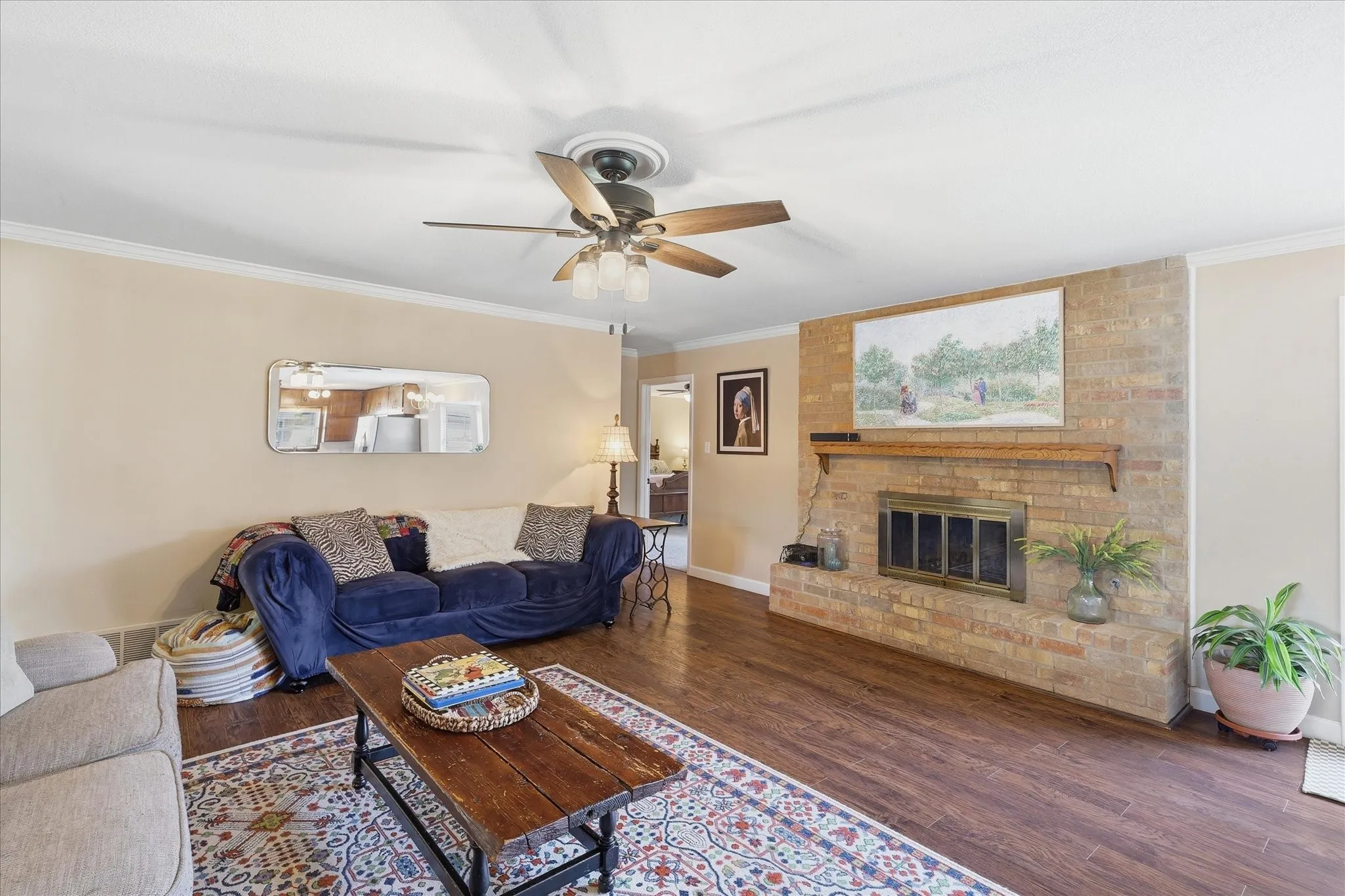 Wood-finish flooring throughout, brick fireplace with a wood mantle, ceiling fan with integrated lighting, and crown molding