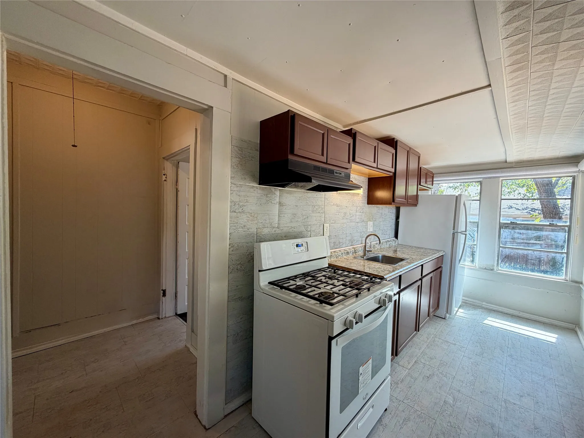 White gas range, stainless steel sink, wood-finish cabinetry, stone-look countertops, and tiled backsplash