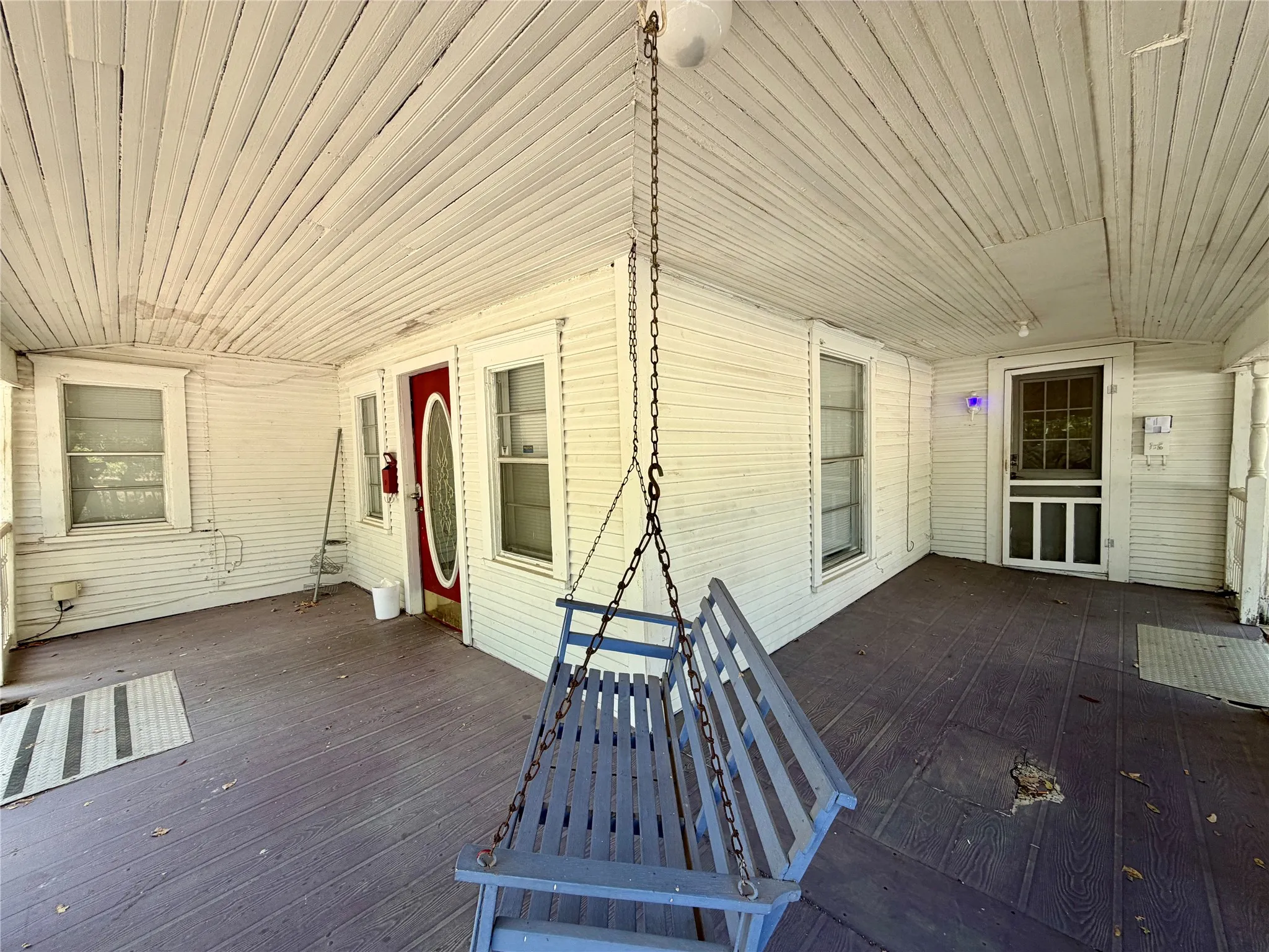 Covered porch with wood panel ceiling and wood flooring, featuring a red entry door with oval glass insert, a second entry door with screen, and multiple windows