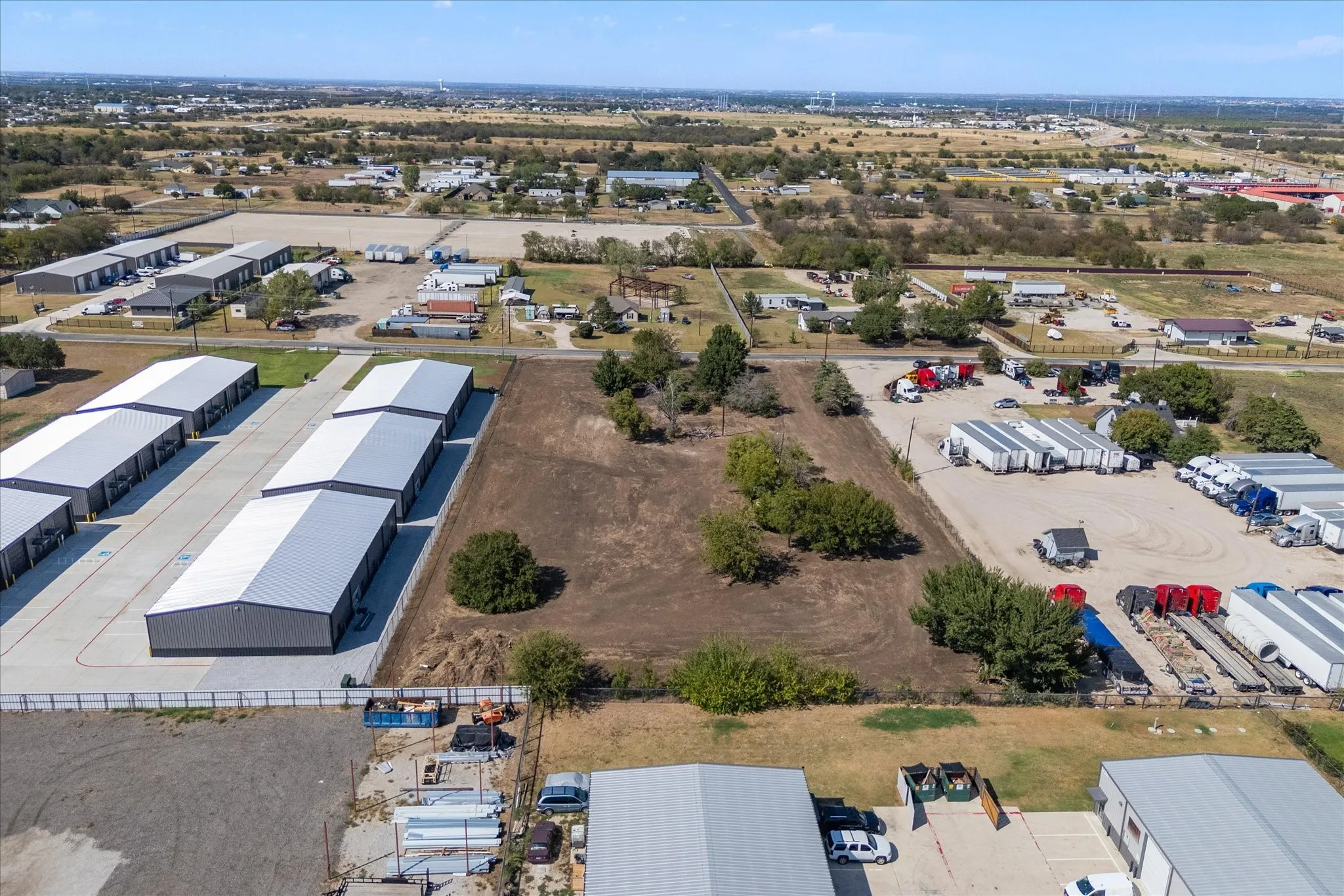 Expansive cleared lot with mature trees, surrounded by commercial structures, and a paved lot