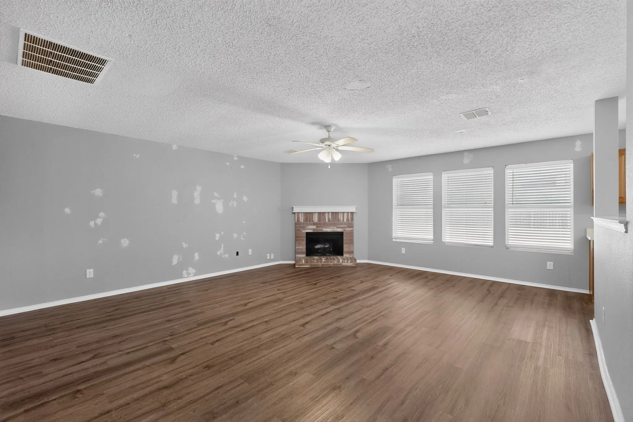 Expansive living area with wood-finish flooring, a brick-surround fireplace, and multiple windows with blinds
