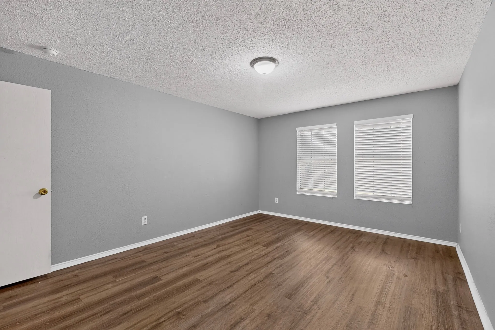 Room featuring wood-finish flooring, light grey walls, white trim, and two windows with horizontal blinds