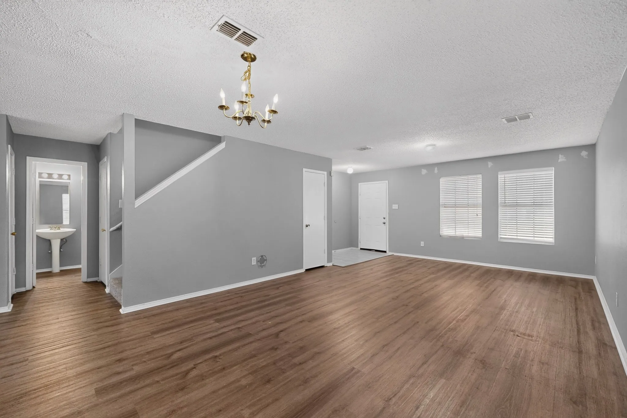 Spacious interior featuring wood-finish flooring, two windows with blinds, a prominent chandelier, and a powder room with a pedestal sink and vanity lighting