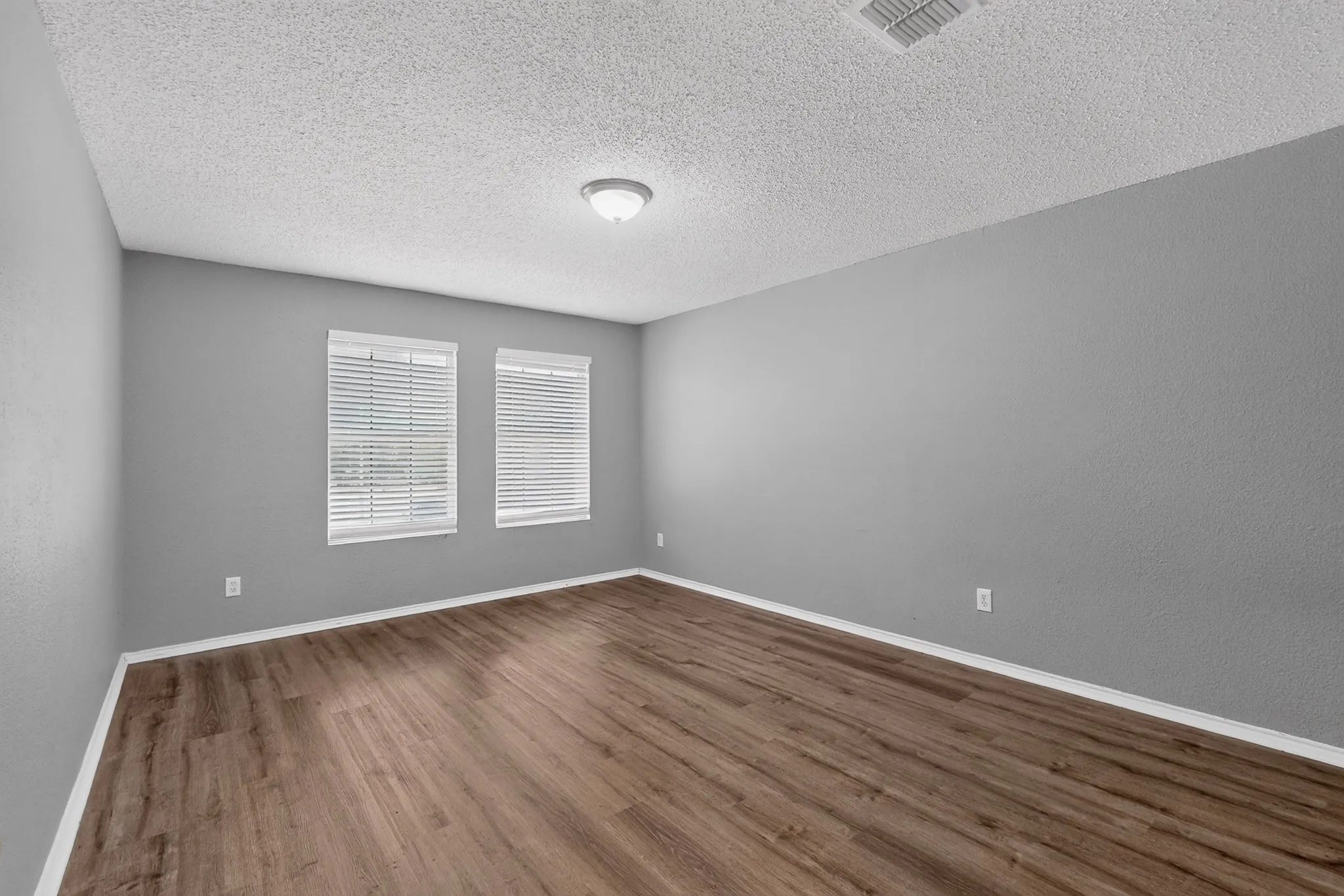 Wood-finish flooring, neutral gray walls, two windows with blinds, a ceiling-mounted light fixture, and textured ceiling