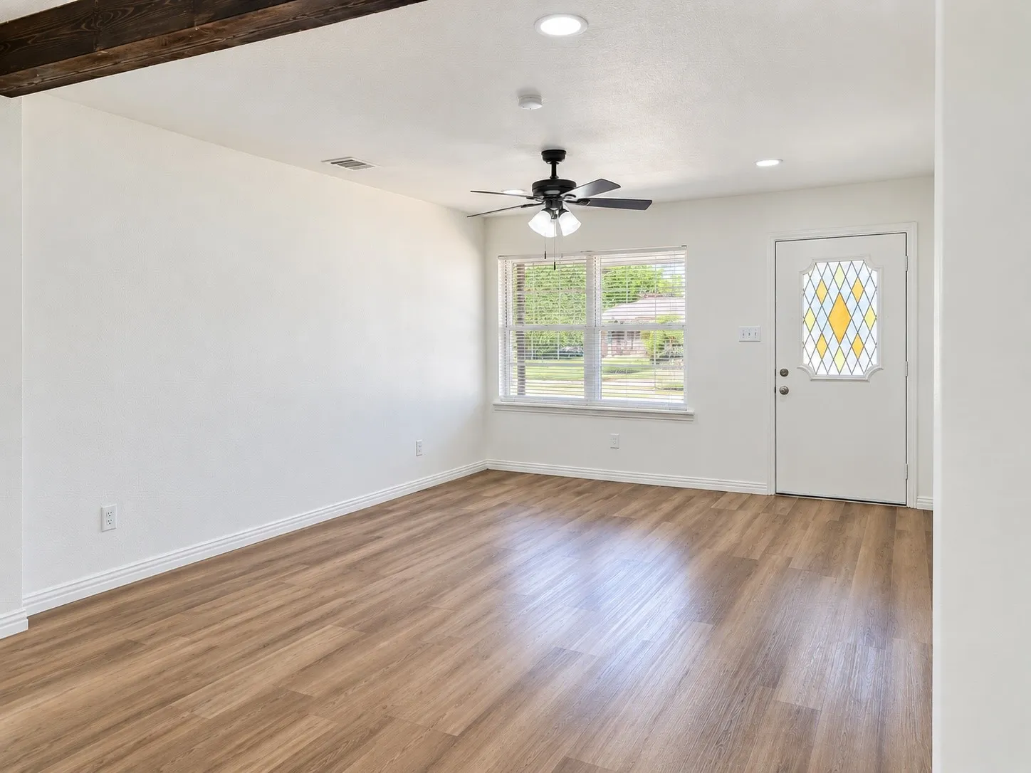 Wood-finish flooring, white walls, exposed wood beam, ceiling fan, and front entry door with leaded glass insert