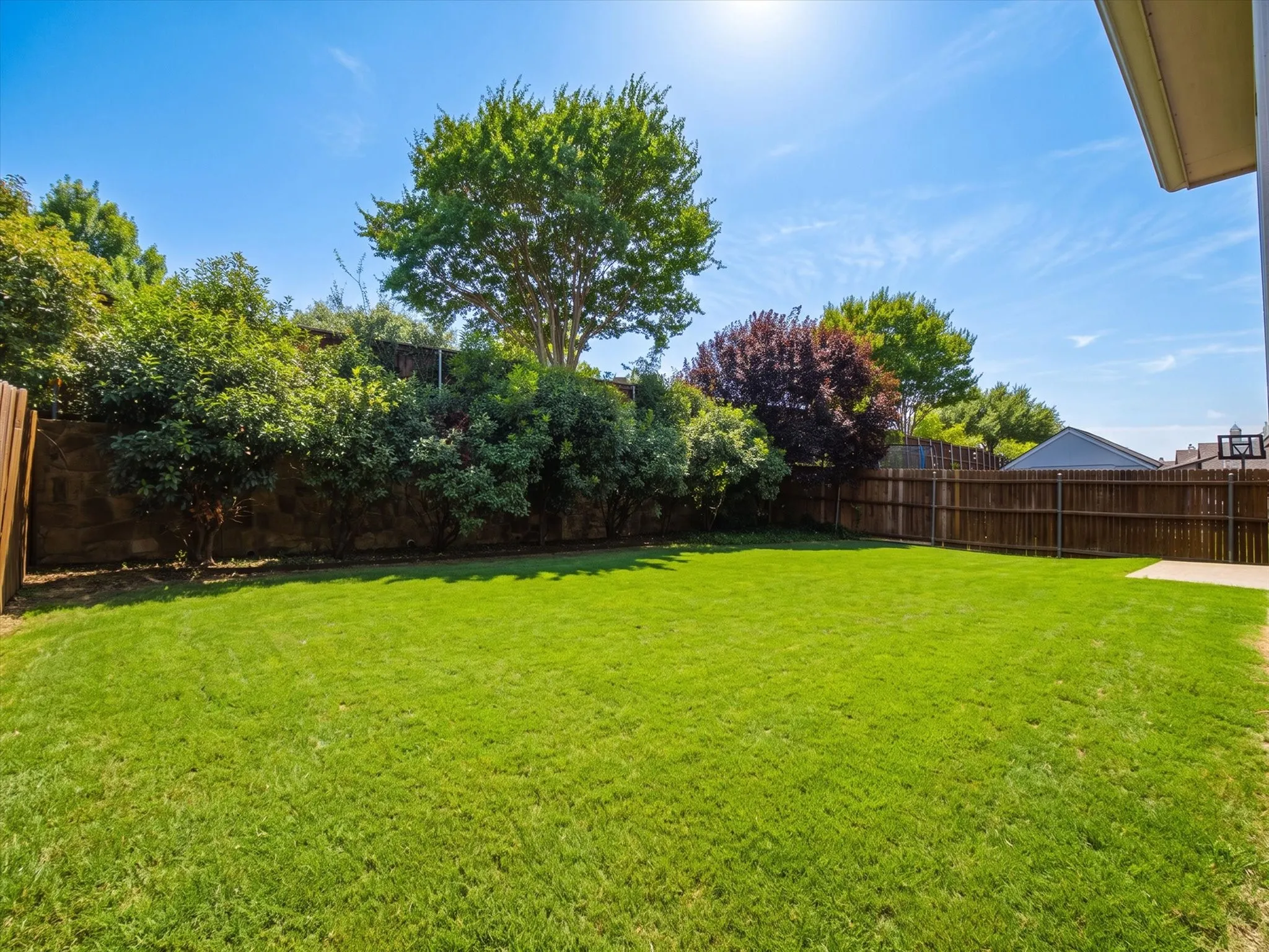 Expansive grassy area bordered by mature trees and shrubs, featuring wood fencing and a concrete patio
