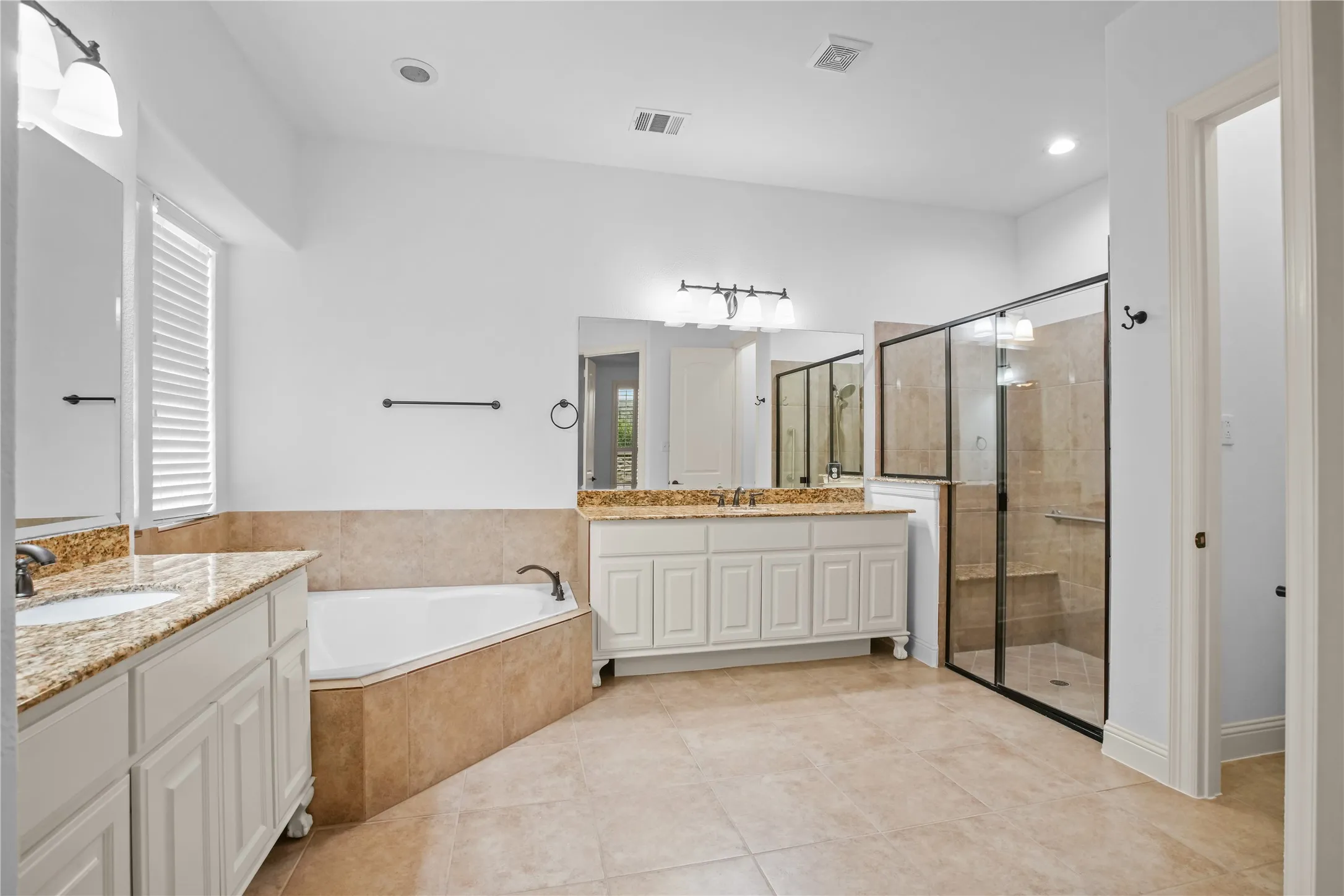 Corner soaking tub, dual vanities with granite countertops, tiled flooring, and a glass-enclosed shower with a built-in bench