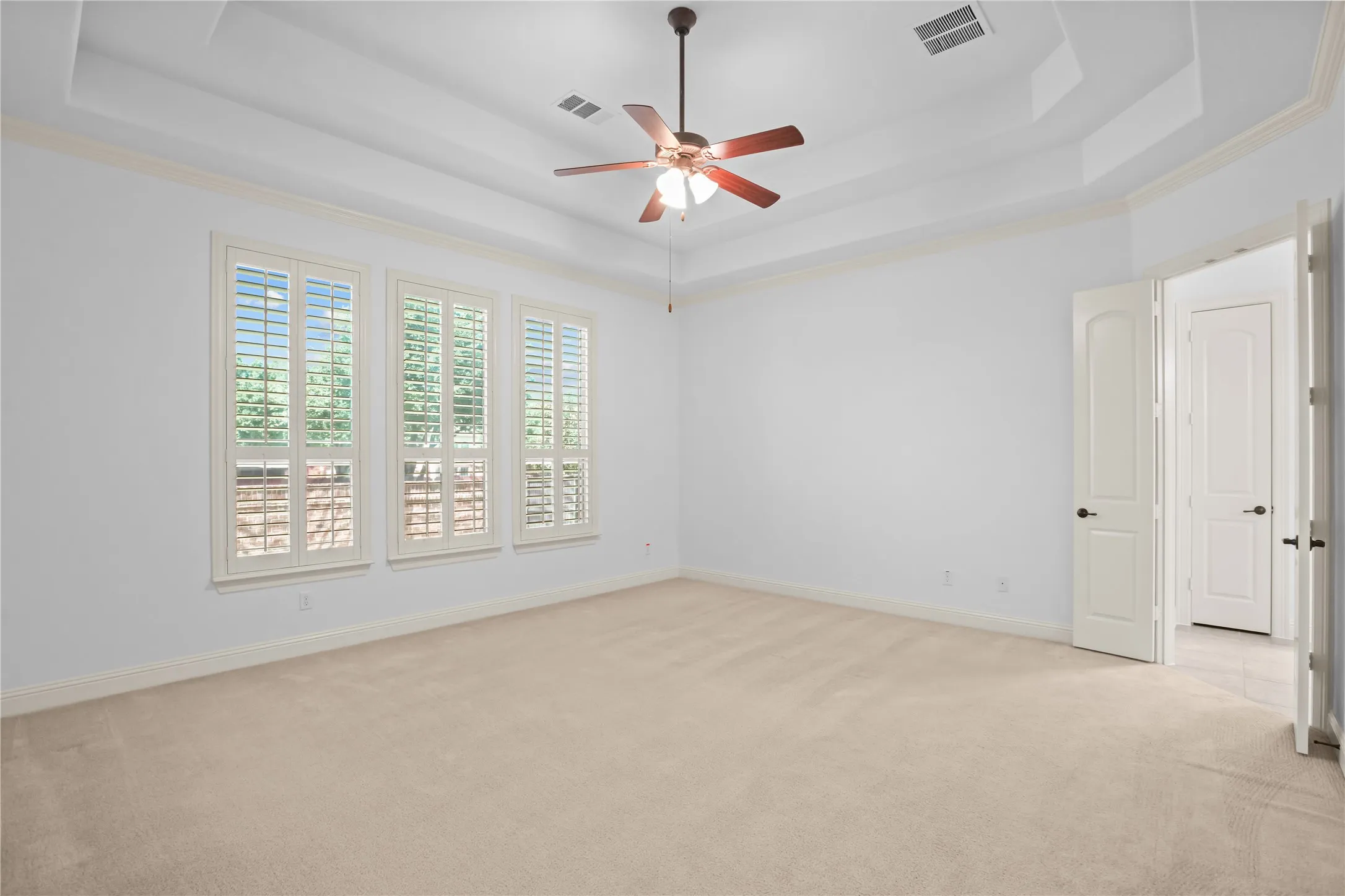 Tray ceiling with a ceiling fan, three windows with plantation shutters, and carpeted flooring