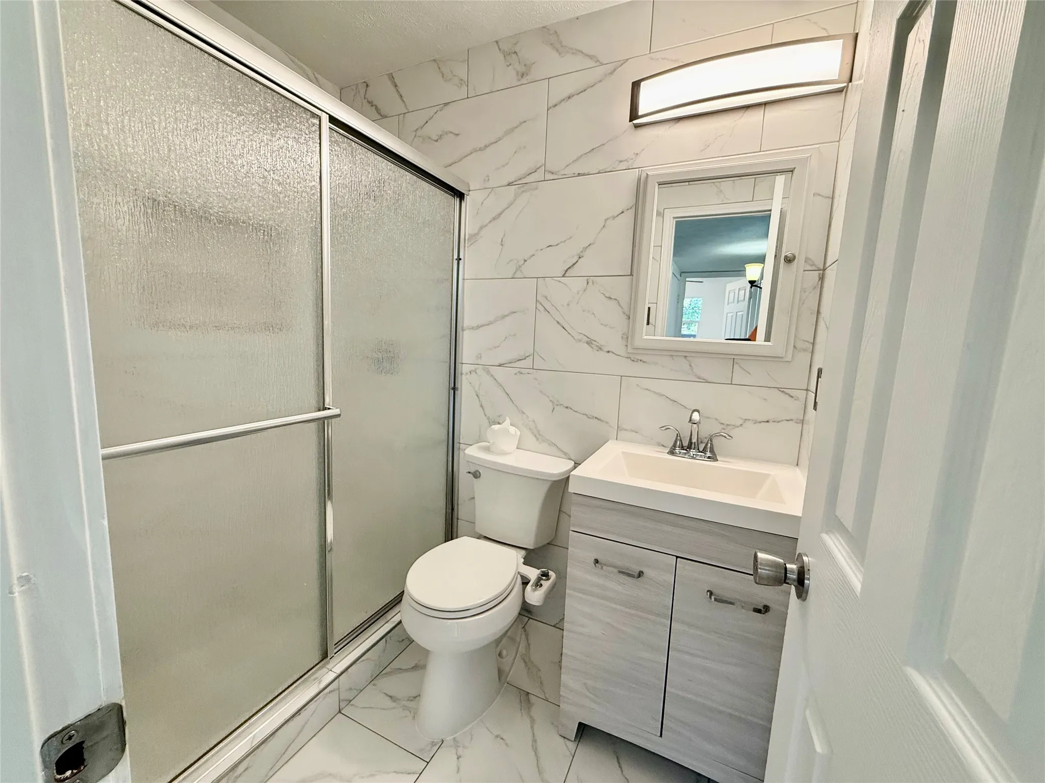 Full-height tiled walls featuring a white marble-look pattern, white vanity with integrated sink, chrome faucet, and a sliding glass shower enclosure