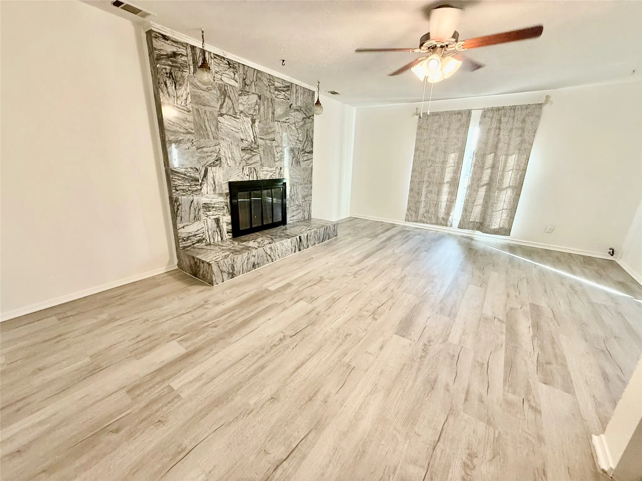 Spacious interior with light wood-finish flooring, a stone-clad fireplace with hearth, and a ceiling fan with integrated lighting